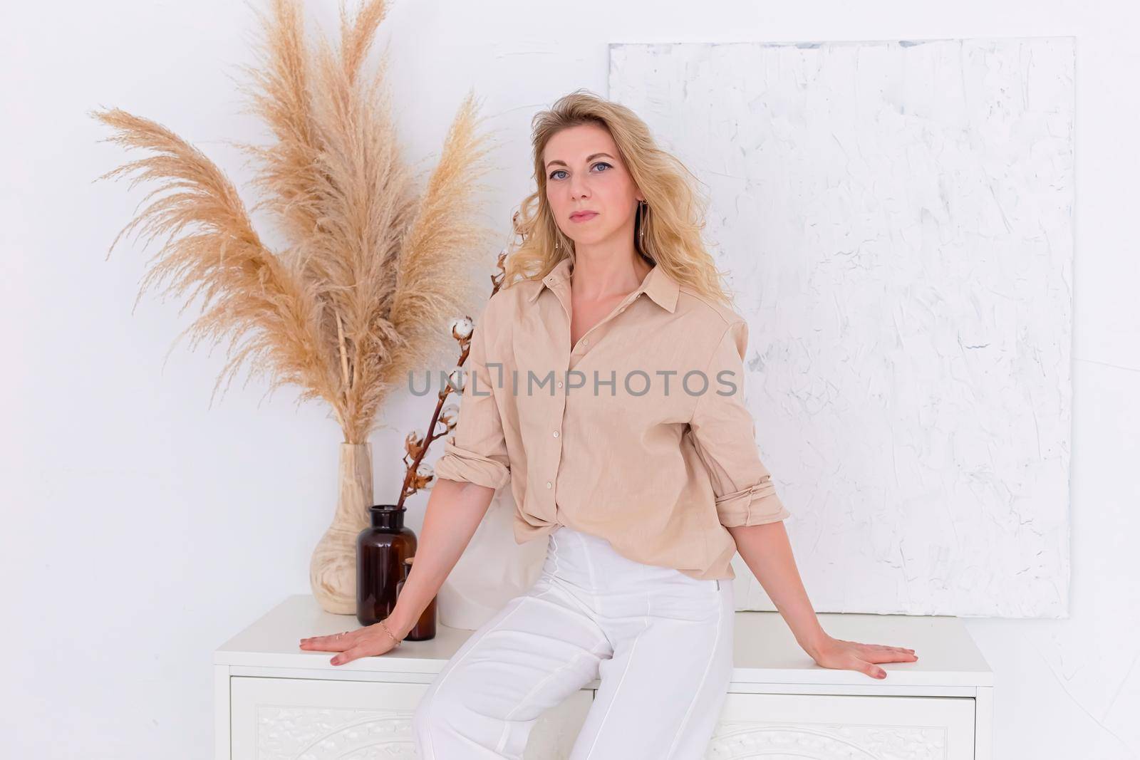 beautiful slender woman 30-40 years old with blond hair in a beige shirt and white trousers in a white room interior sits on a white chest of drawers against the wall, looks at the camera. Copy space.