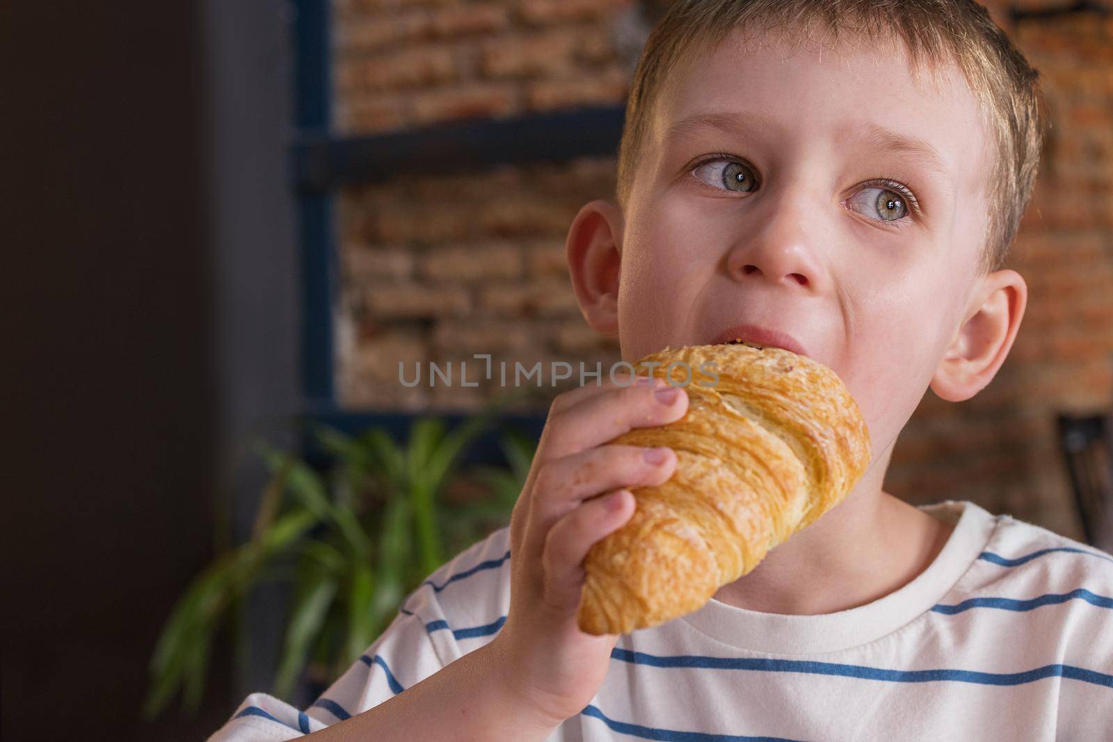 Portrait of boy eating a croissant in a cafe, close-up. copy space for your text by Ramanouskaya