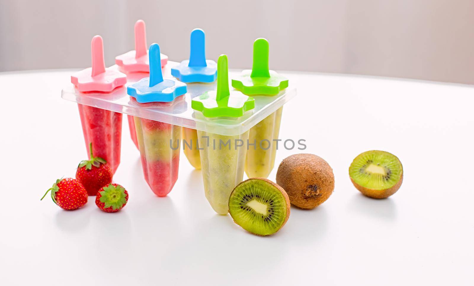 A plastic mold with multi-colored homemade kiwi and strawberry ice cream stands on a white table by the window in daylight. Near kiwi and strawberry