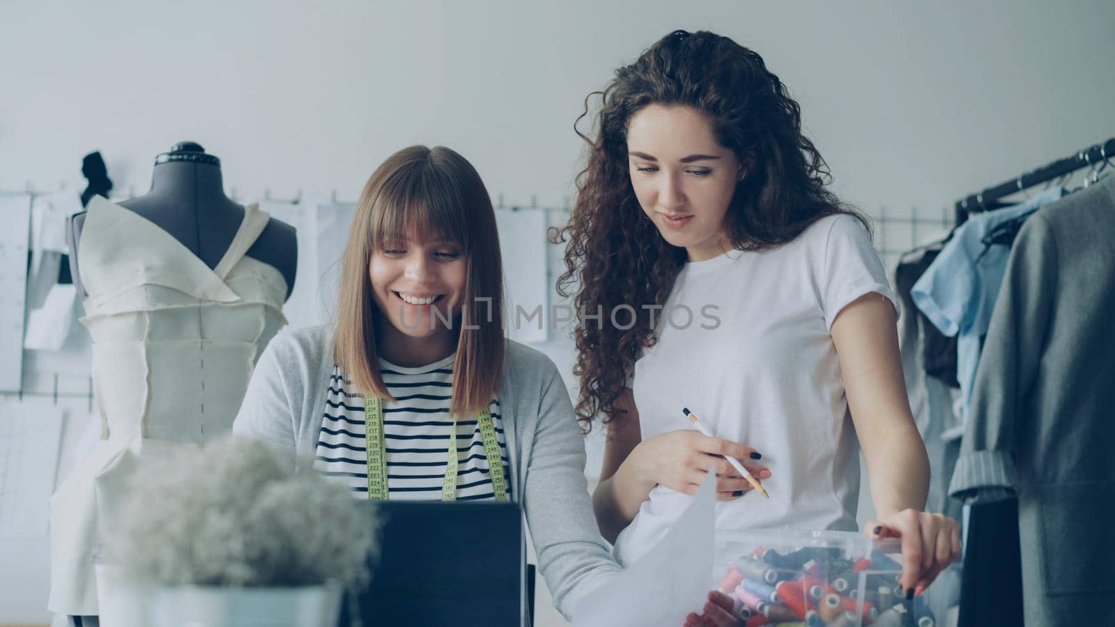 Cheerful creative clothing designers are watching laptop screen, having friendly conversation, smiling and laughing together in light workshop. Fun at workplace concept. by silverkblack