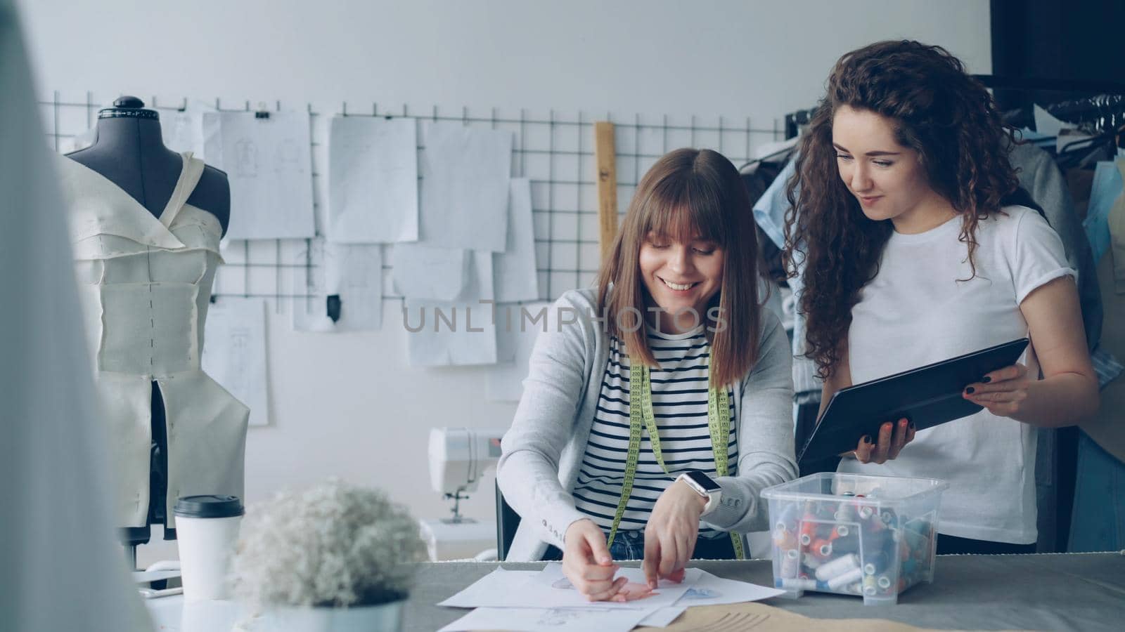 Female employee of tailor shop is measuring garment drawings with sewing threads while her attractive colleague is showing her tablet screen and talking to her.