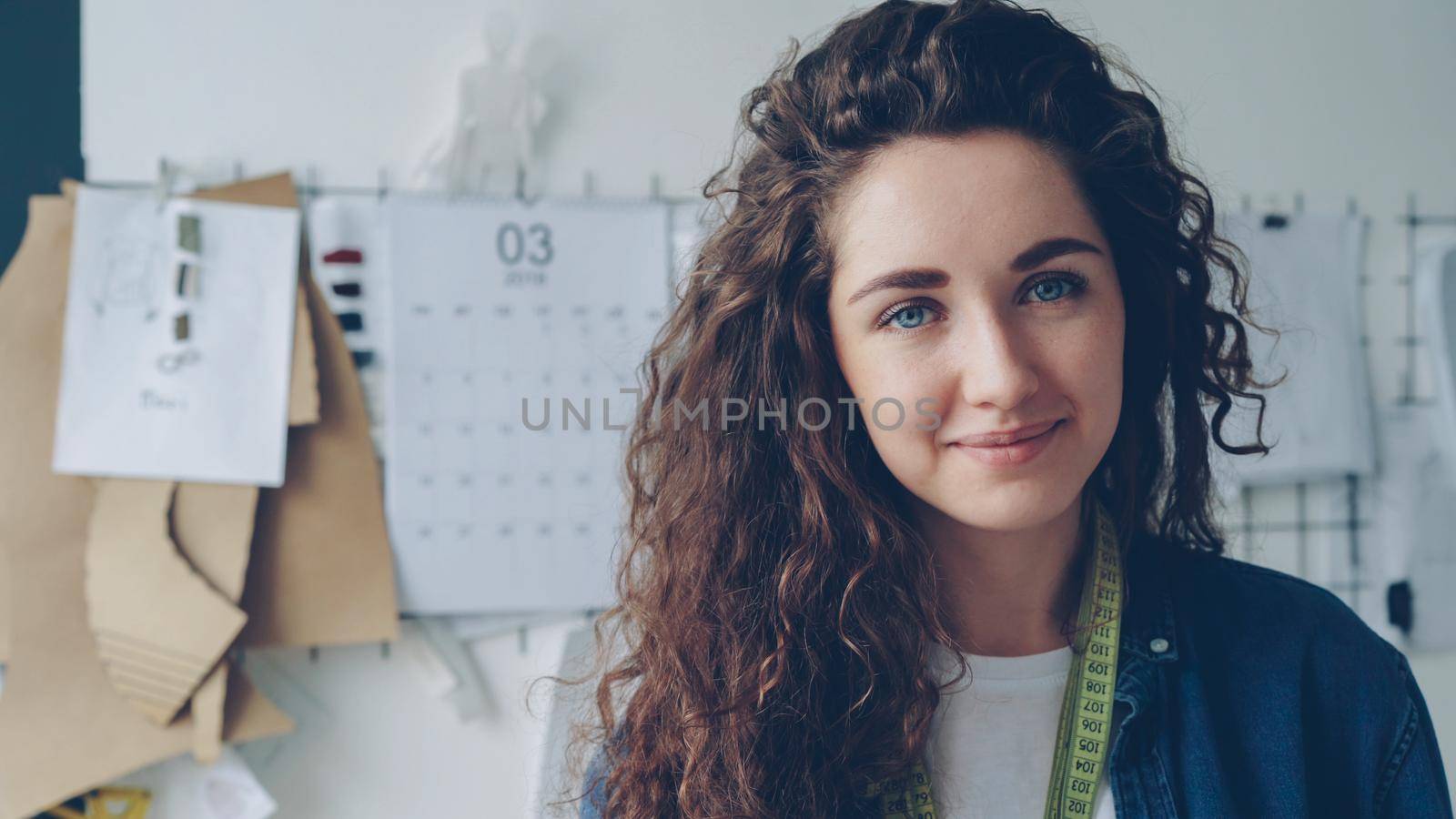 Close-up portrait of young woman seamstress looking at camera in light studio with garment sketches hanging on wall in background. Pretty expressive emotional face, long hair. by silverkblack