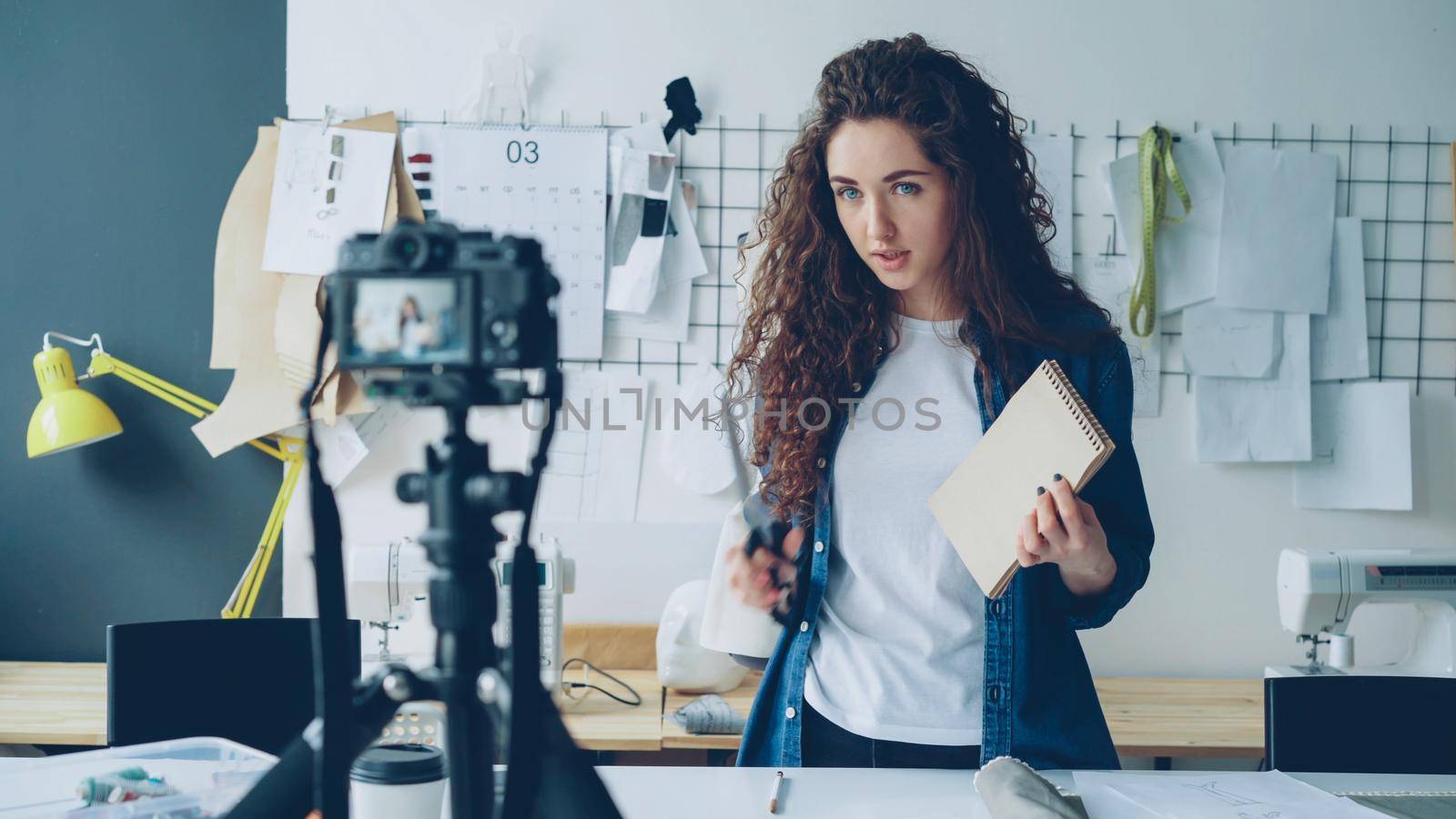 Young attractive seamstress fashion blogger is recording video with camera, talking and showing sketches and tailoring items. Blogging for successful startup concept. by silverkblack