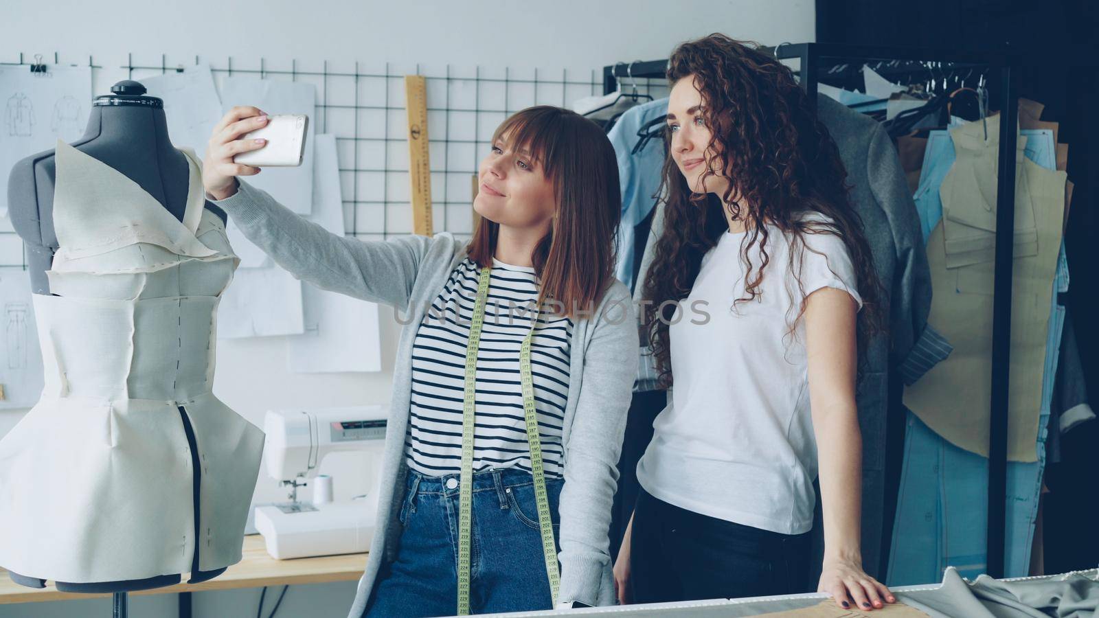 Two cheerful female clothing designers are making funny selfie with smart phone while standing beside clothed mannequin in studio. Attractive women are smiling and posing.