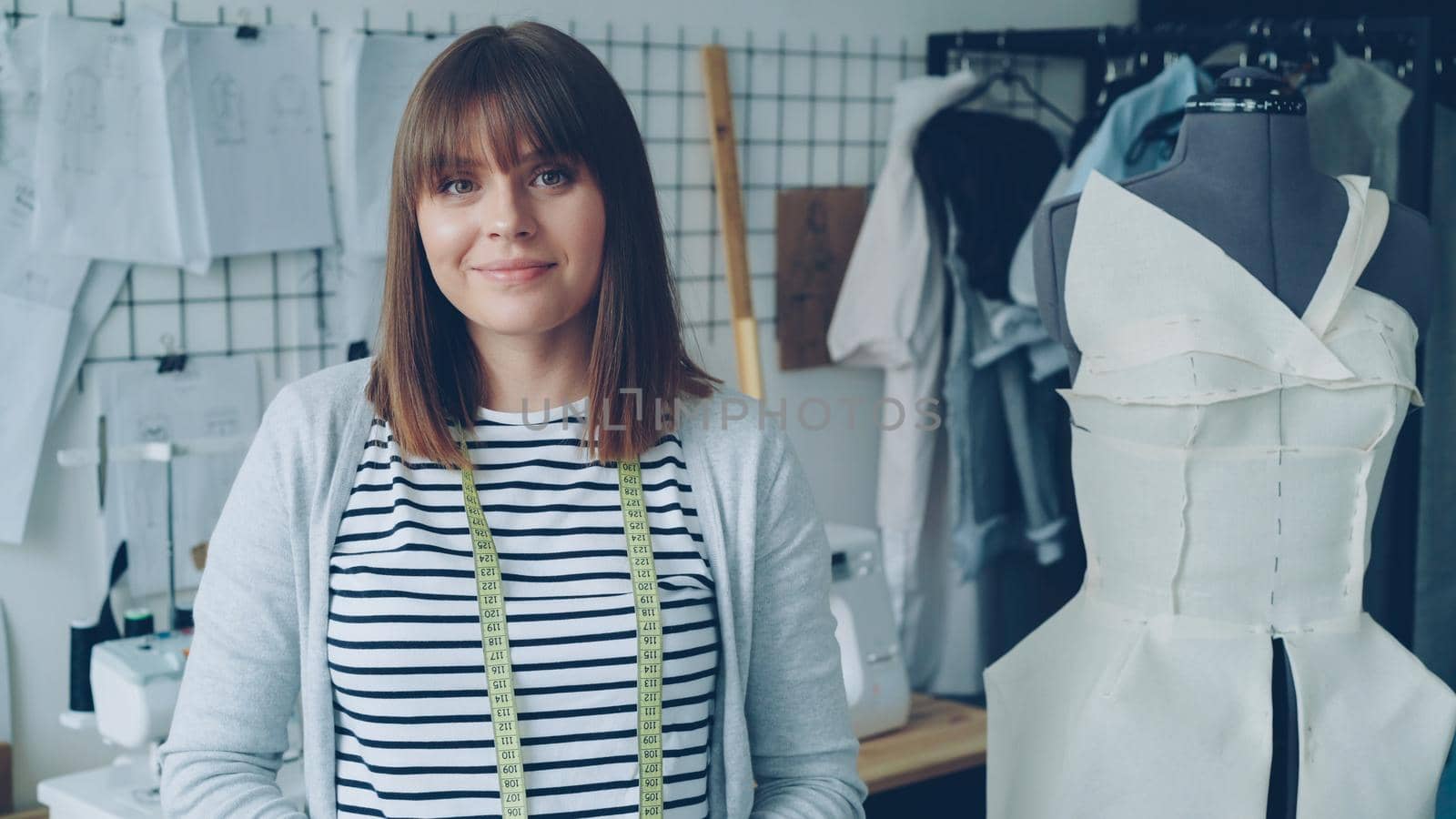 Portrait of attractive young woman clothing designer with brown hair looking at camera. Smiling woman is standing beside dummy, light modern tailoring workshop in background. by silverkblack