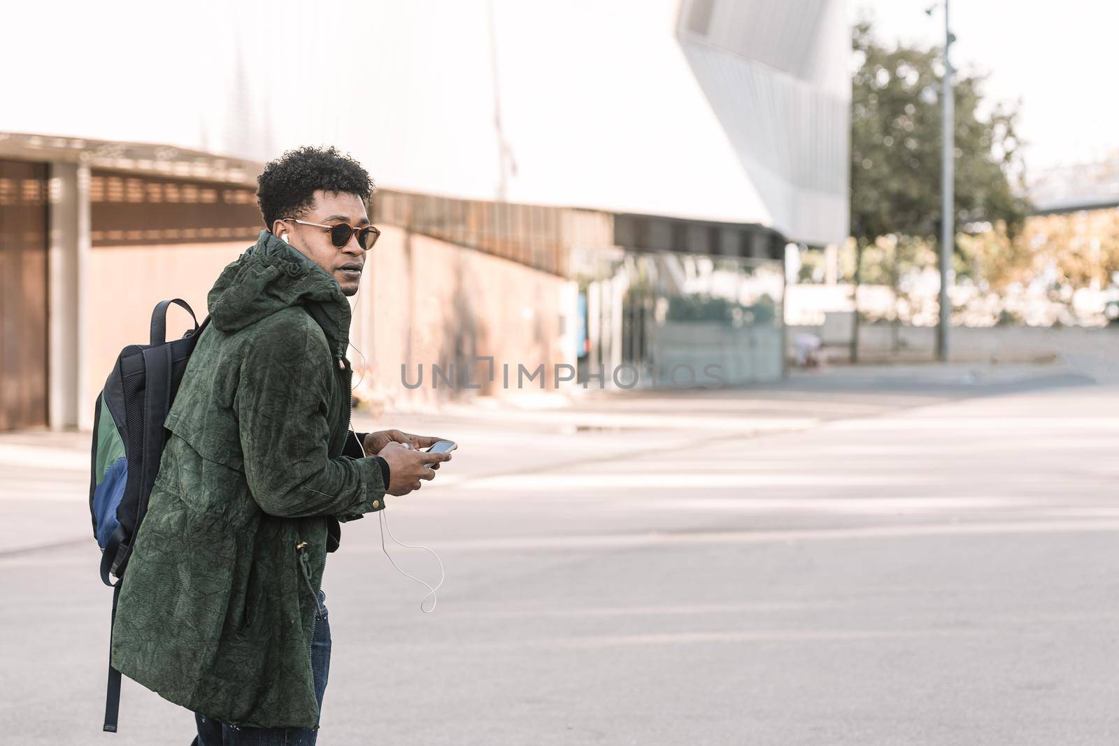 african american young male with earphones walking by the city and listening music with a mobile phone, technology and lifestyle concept using internet electronic device outdoors