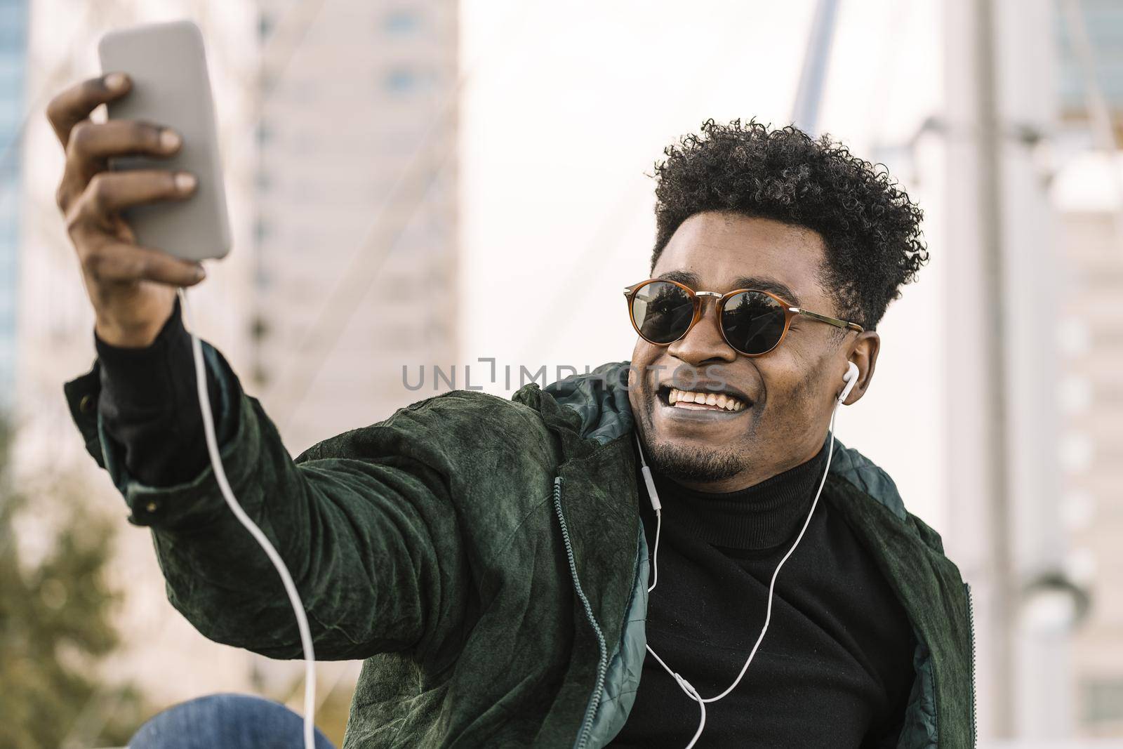 handsome young african black man with sunglasses making a video call with smartphone and earphones while sitting in the city, lifestyle and technology concept using internet electronic device