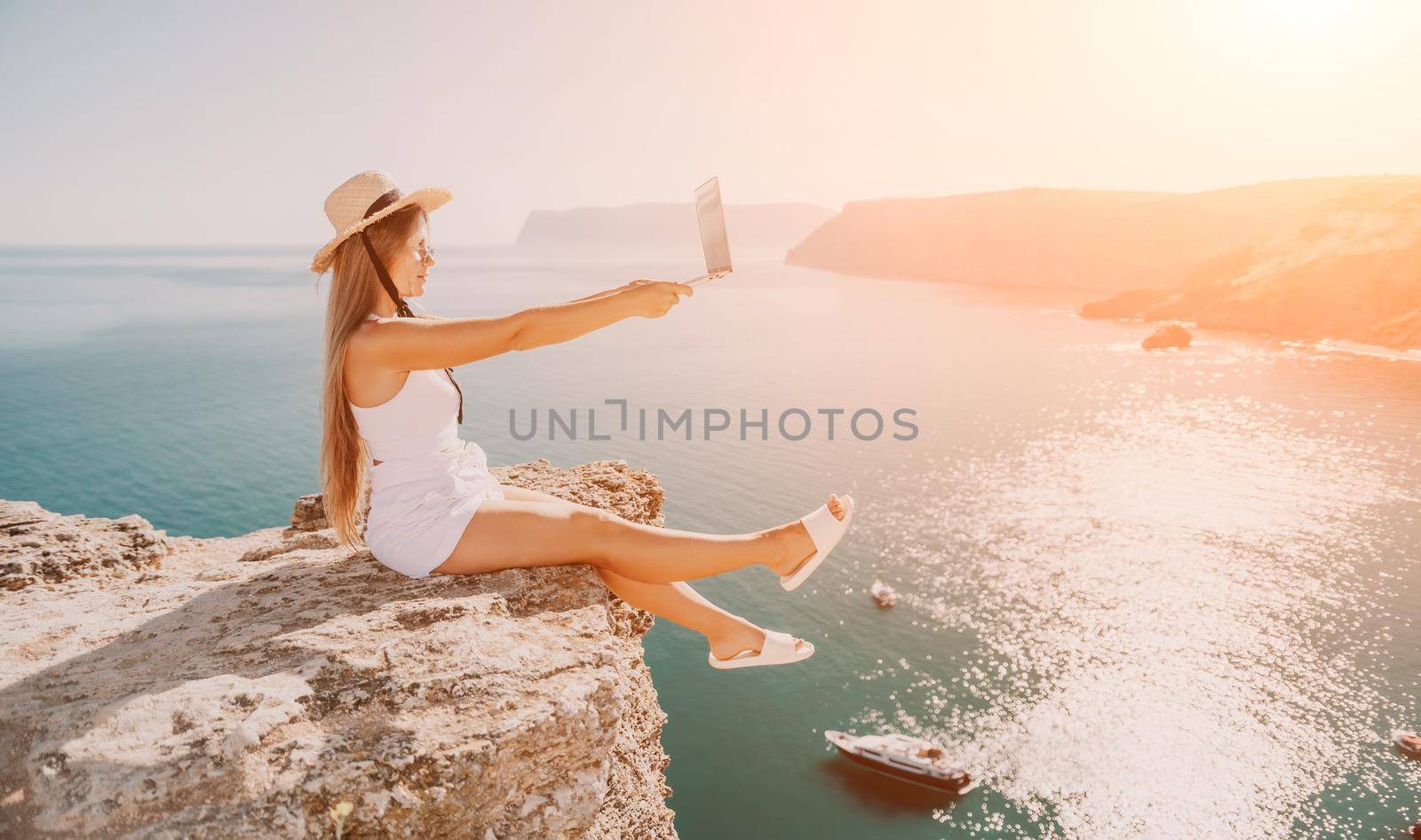 Successful business woman in yellow hat working on laptop by the sea. Pretty lady typing on computer at summer day outdoors. Freelance, travel and holidays concept.