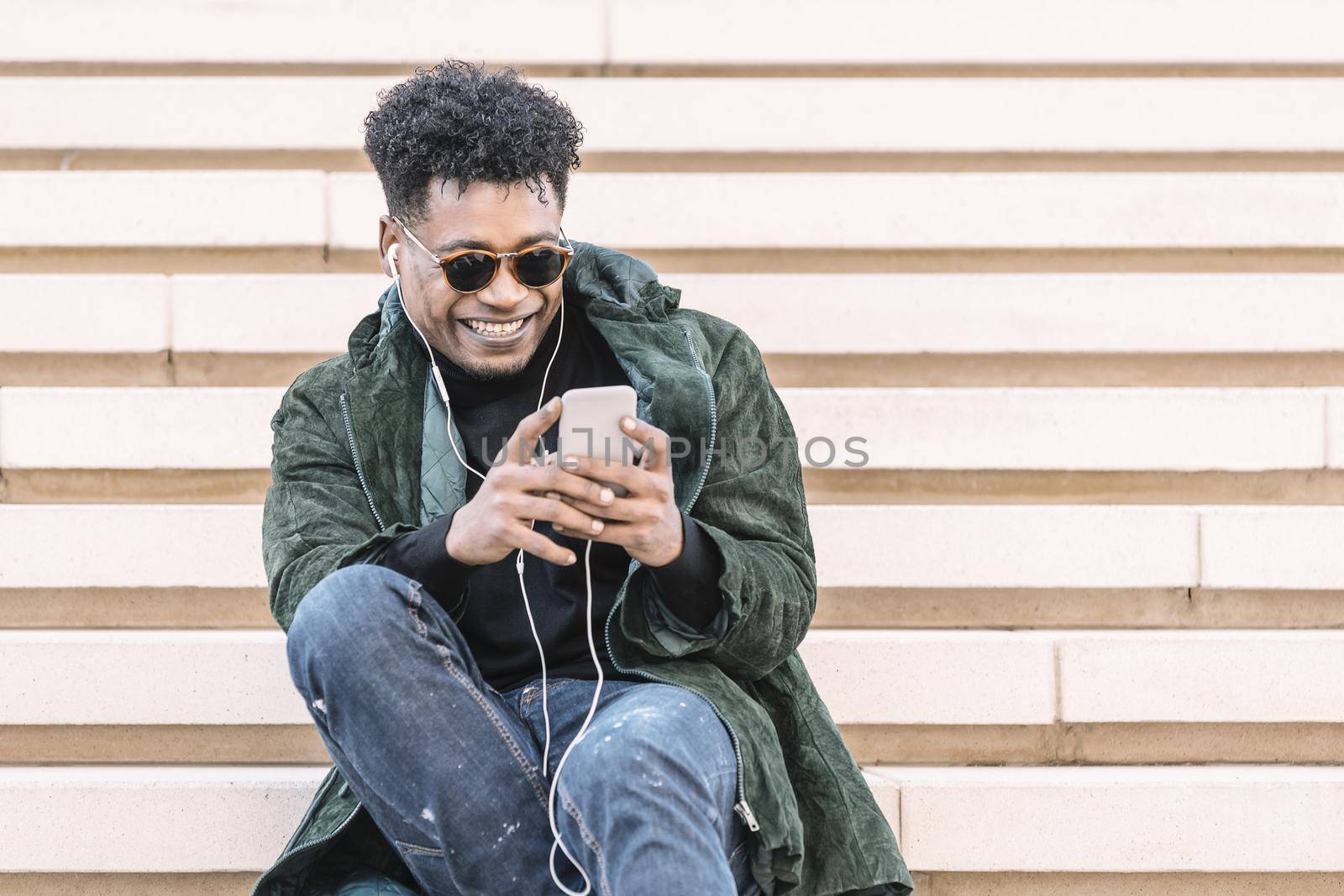 african american young male in casual modern clothes with phone smiling and sitting on stairs while listening to music in earphones, technology and lifestyle concept