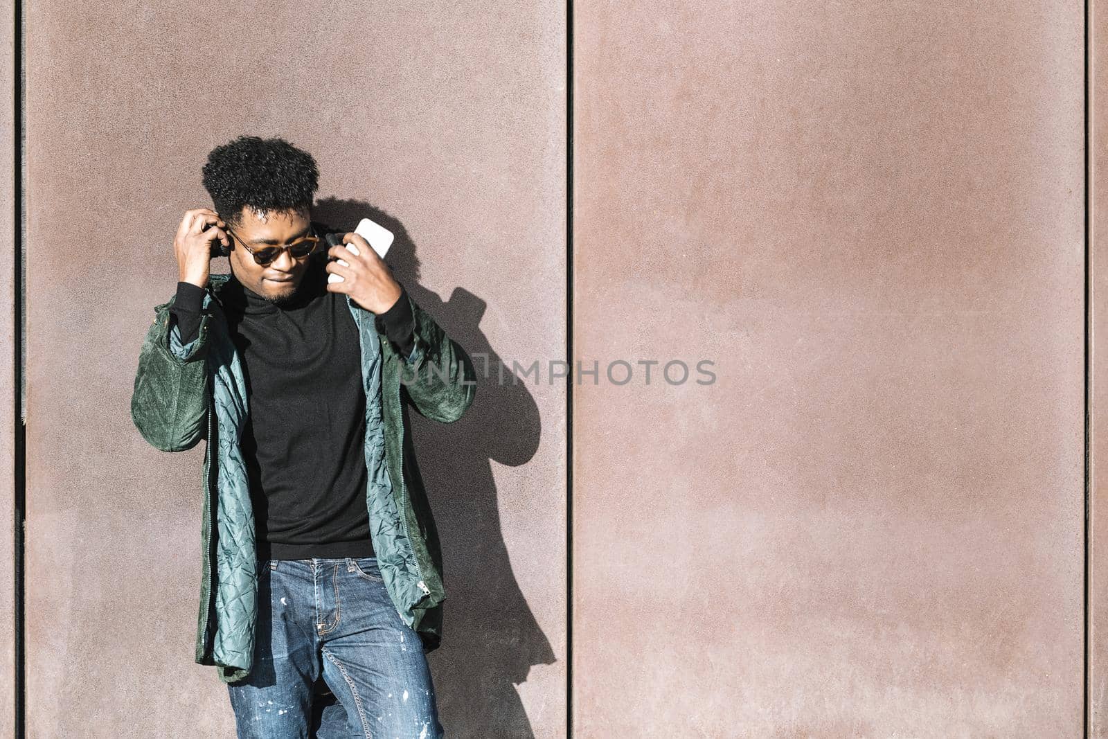 cool african black man with casual modern clothes and a mobile phone and headphones listening music leaning against wall, technology and lifestyle concept