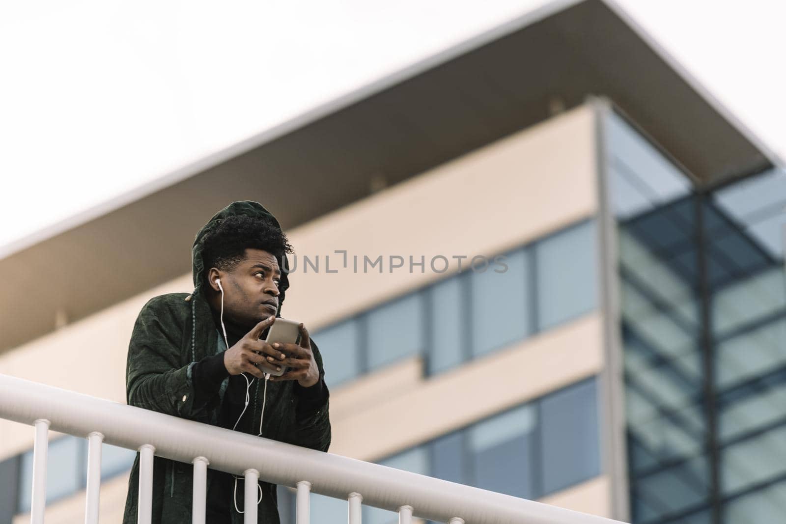 portrait of a smiling young attractive african black man listening to music with earphones and mobile phone while leaning on a handrail outdoors in the city, technology and lifestyle concept