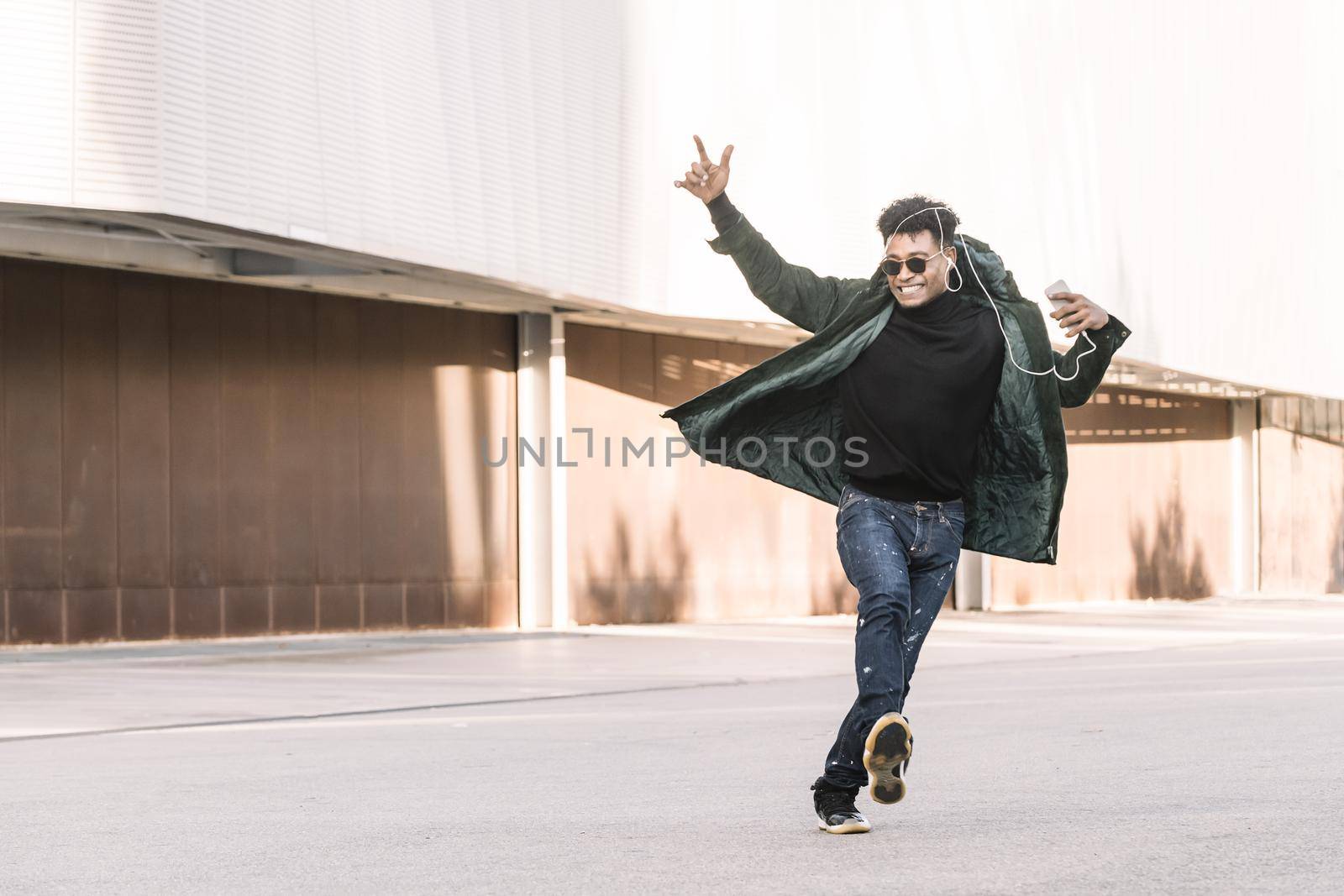 black young man with earphones dancing happy by the city and listening music with a mobile phone, technology and lifestyle concept using internet electronic device outdoors