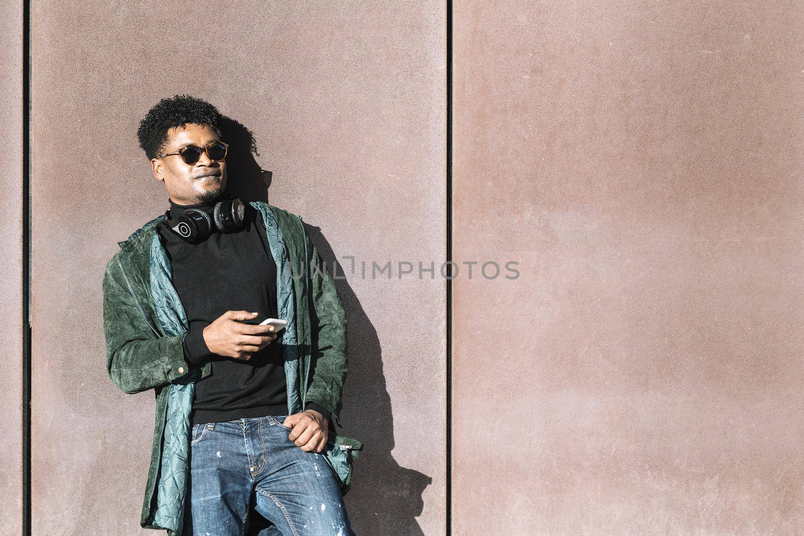 cool african black guy with casual modern clothes and a mobile phone and headphones to listen music leaning against wall, technology and lifestyle concept