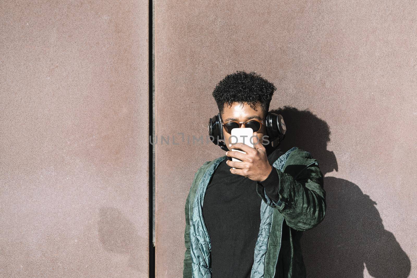 cool african black man with casual modern clothes and headphones to listen music taking a selfie with the phone leaning against wall, technology and lifestyle concept