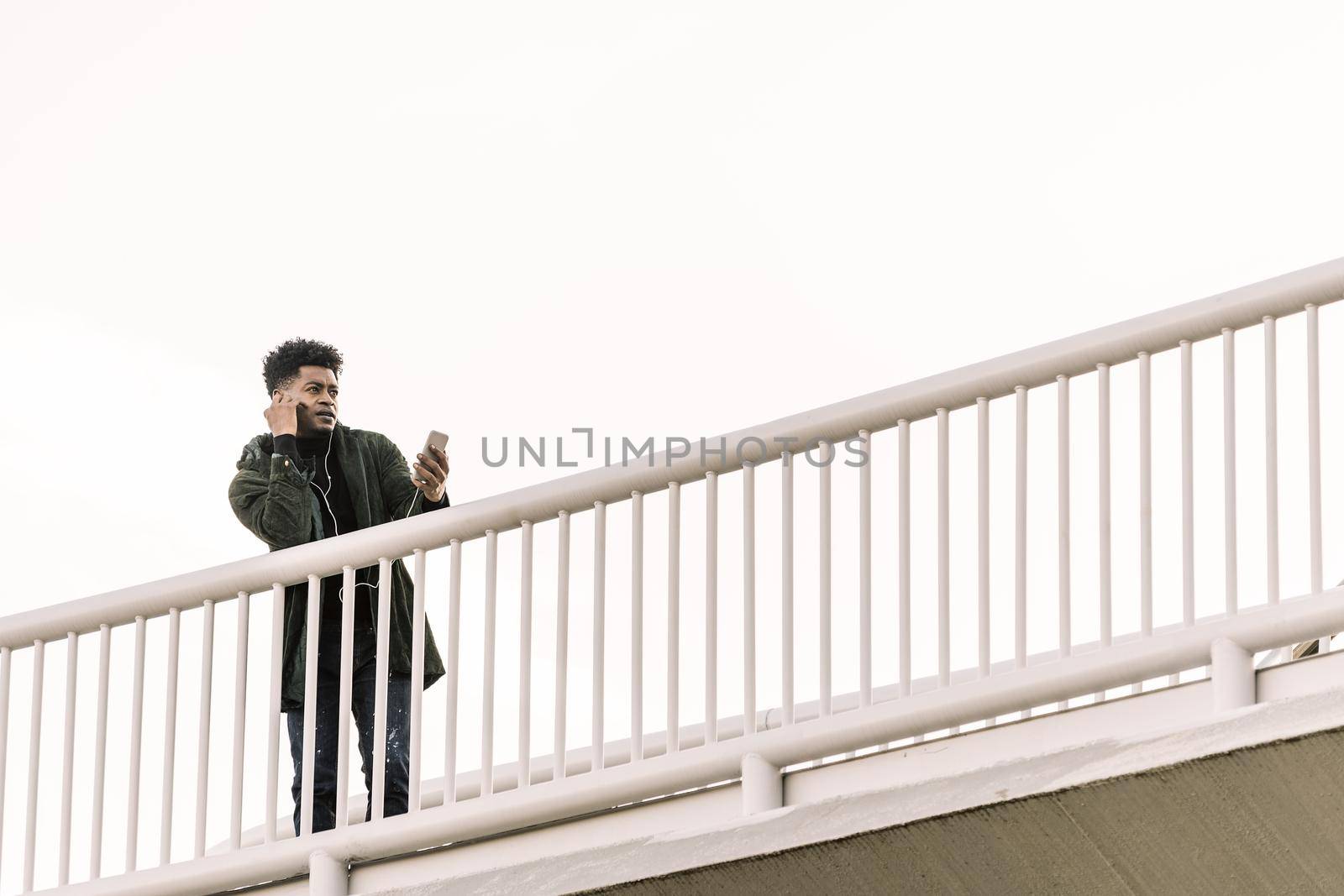 african black man with headphones making video call with a mobile phone leaning on a handrail outdoors in the city, technology and lifestyle concept using internet electronic device