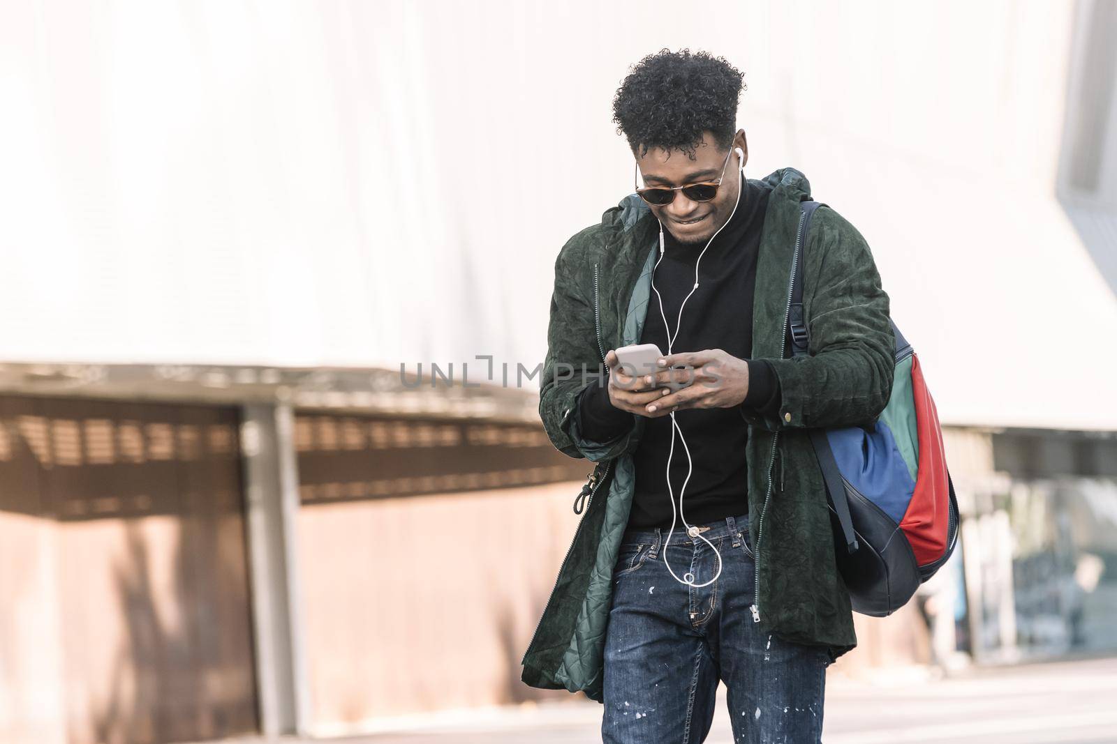 black young man with earphones walking by the city and texting with a mobile phone, technology and lifestyle concept using internet electronic device outdoors