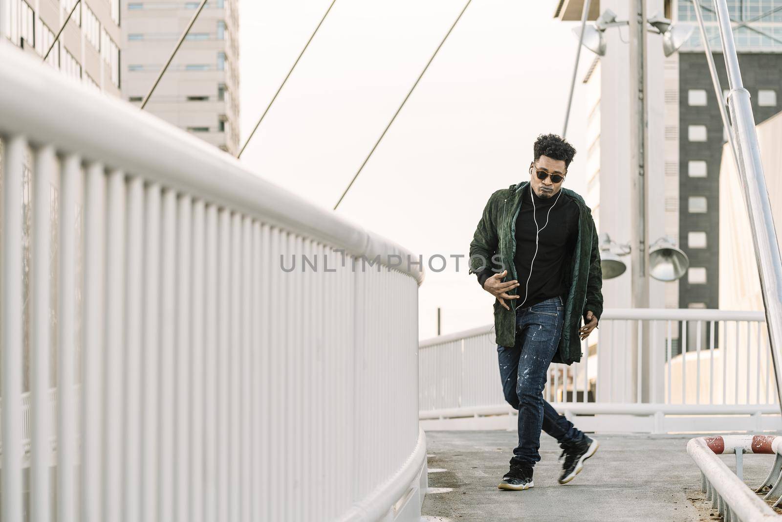 young stylish black man with white earphones and sun glasses dancing outdoor in the city having some fun, lifestyle and technology concept, copy space for text
