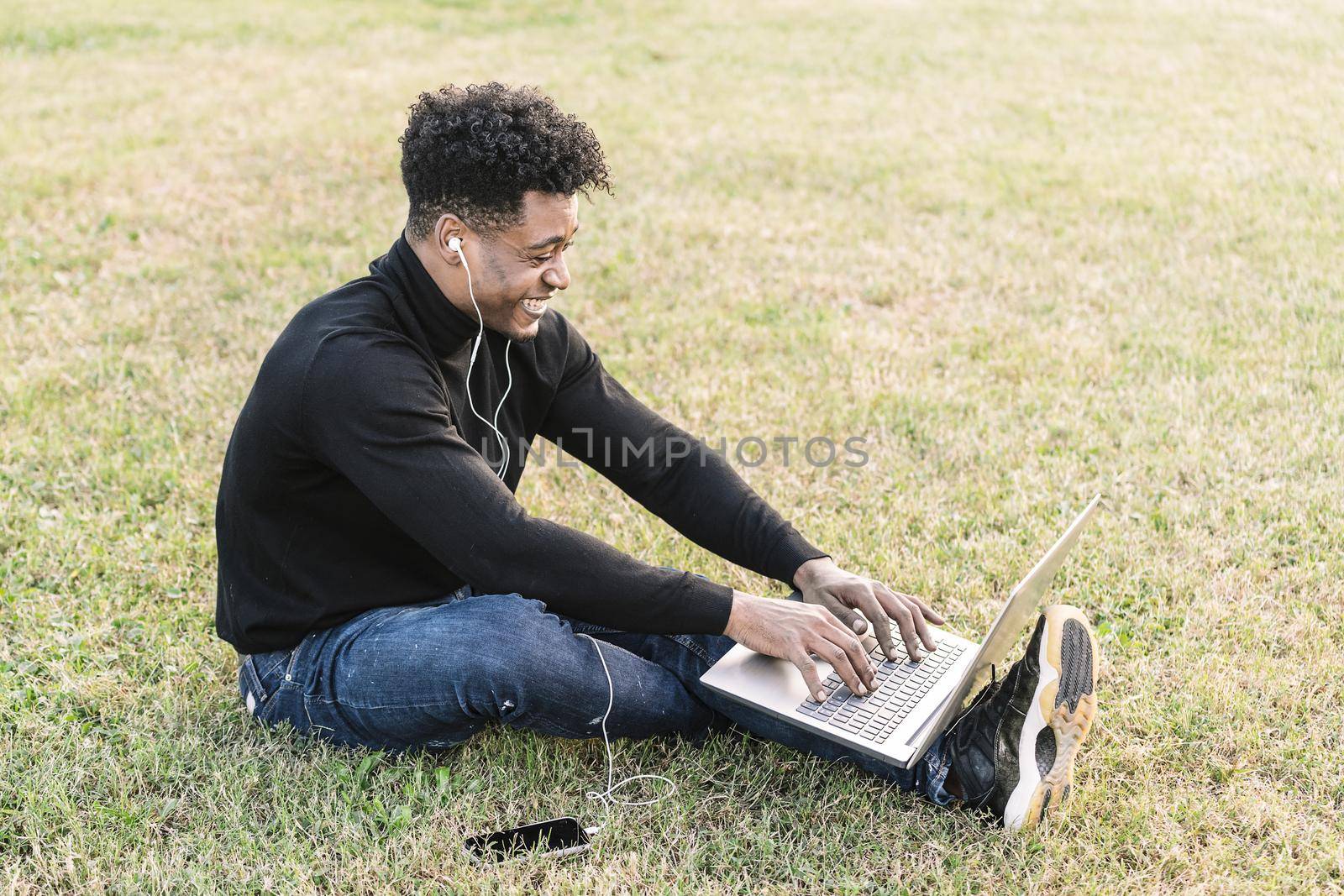 smiling young black student laughing with white earphones and phone working typing in a laptop sitting on the lawn at park, lifestyle and technology concept using internet electronic device