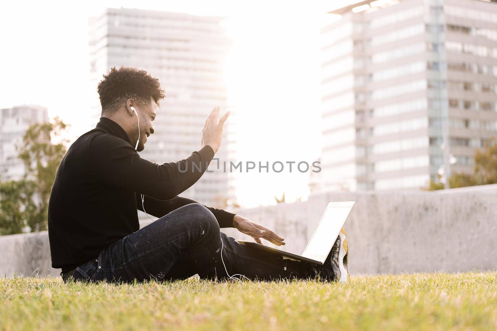smiling young black man laughing with white earphones making a video call with the laptop sitting on the lawn in the city at sunset, lifestyle and technology concept using internet electronic device