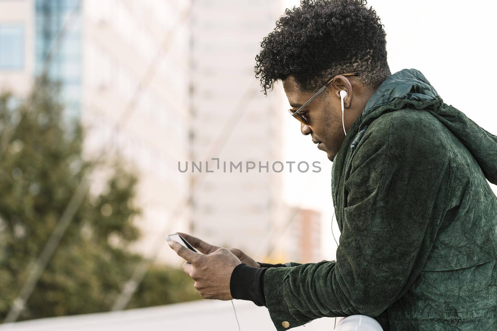 cool young african black man with sunglasses and earphones texting message on smartphone while leaning on a handrail outdoors in the city, lifestyle and technology concept, copy space for text