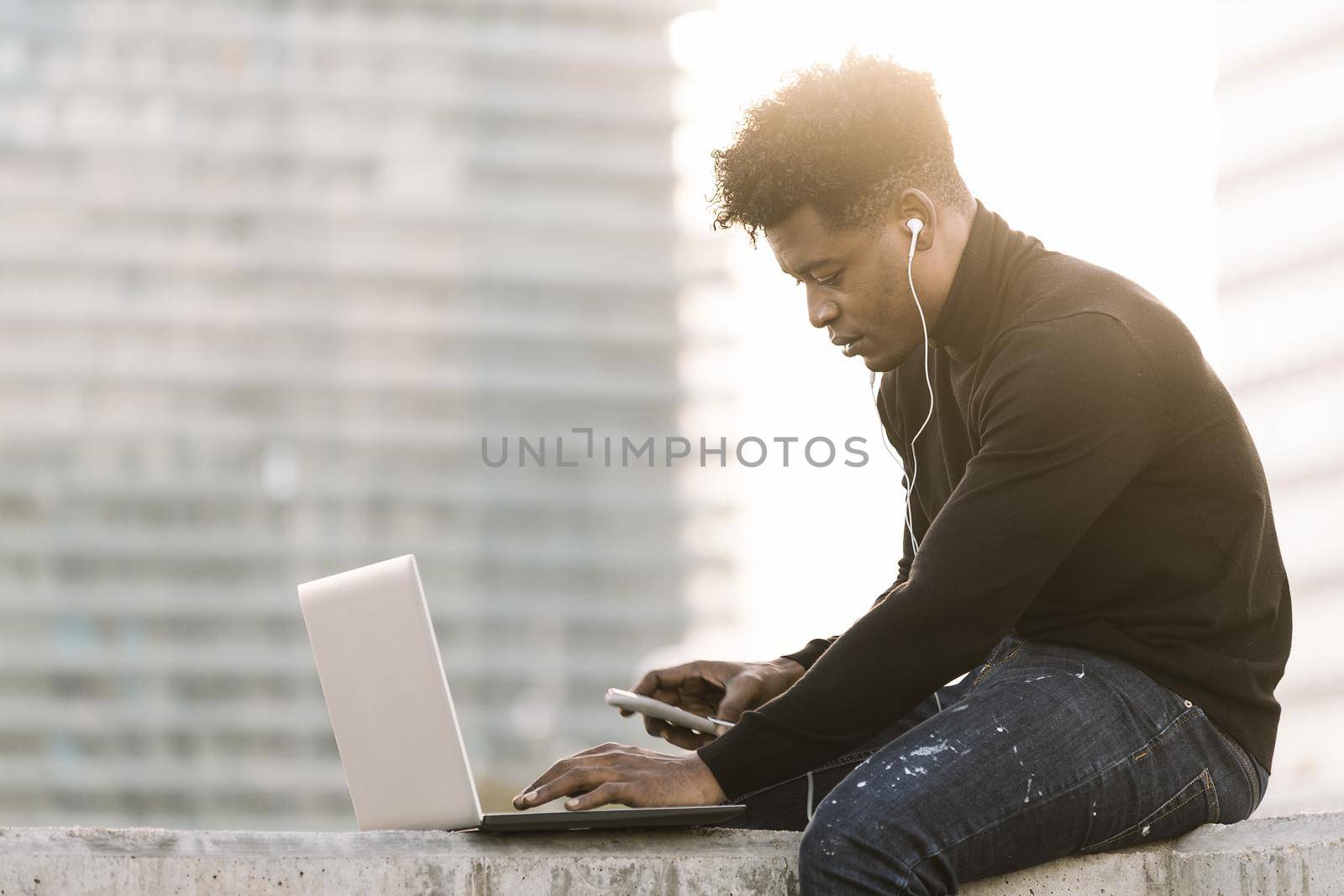 handsome black man with white earphones working with computer and phone sitting outdoors in the city at sunset, lifestyle and technology concept using internet electronic device