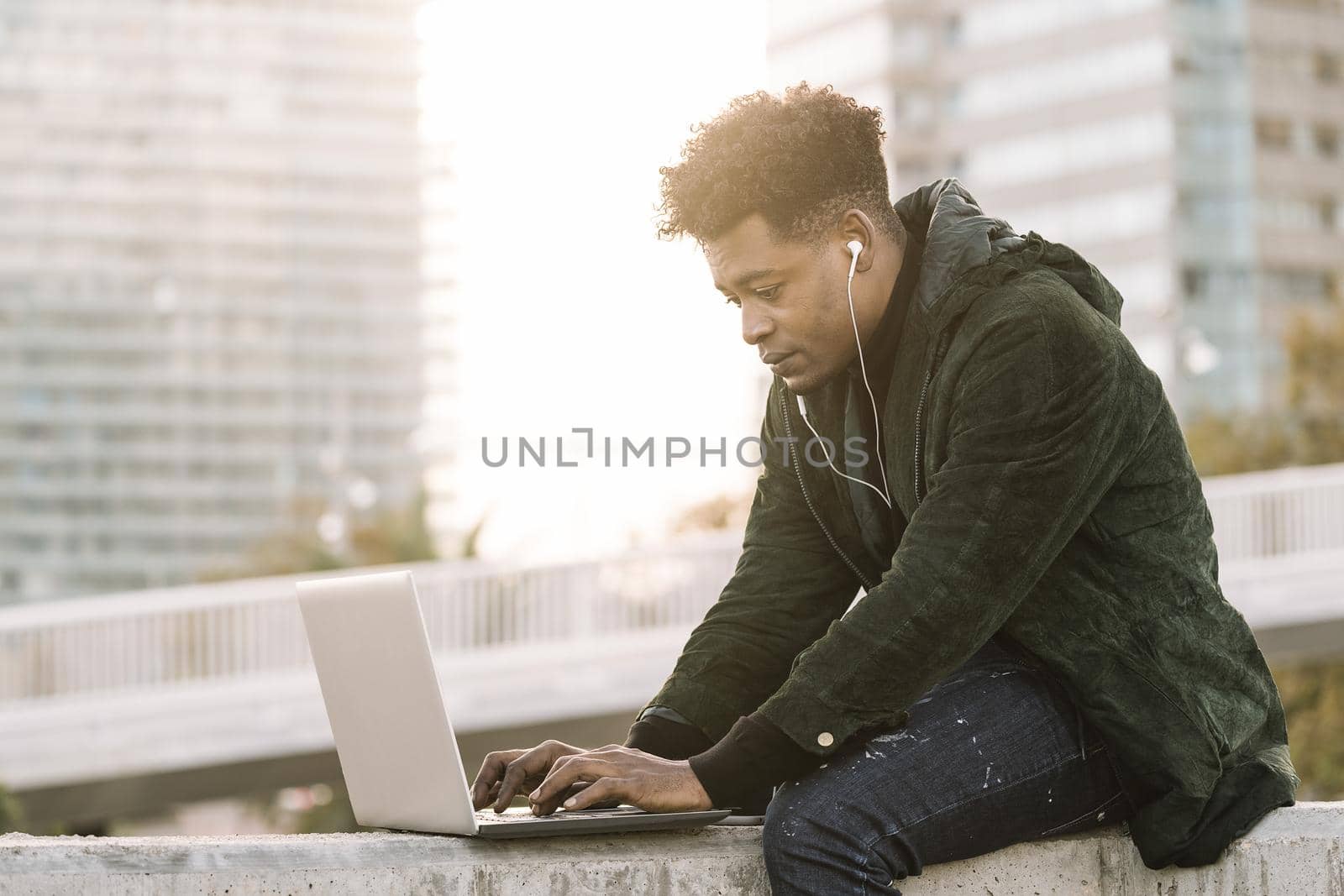 handsome young student with white earphones working typing in a laptop sitting outdoors in the city at sunset, lifestyle and technology concept using internet electronic device