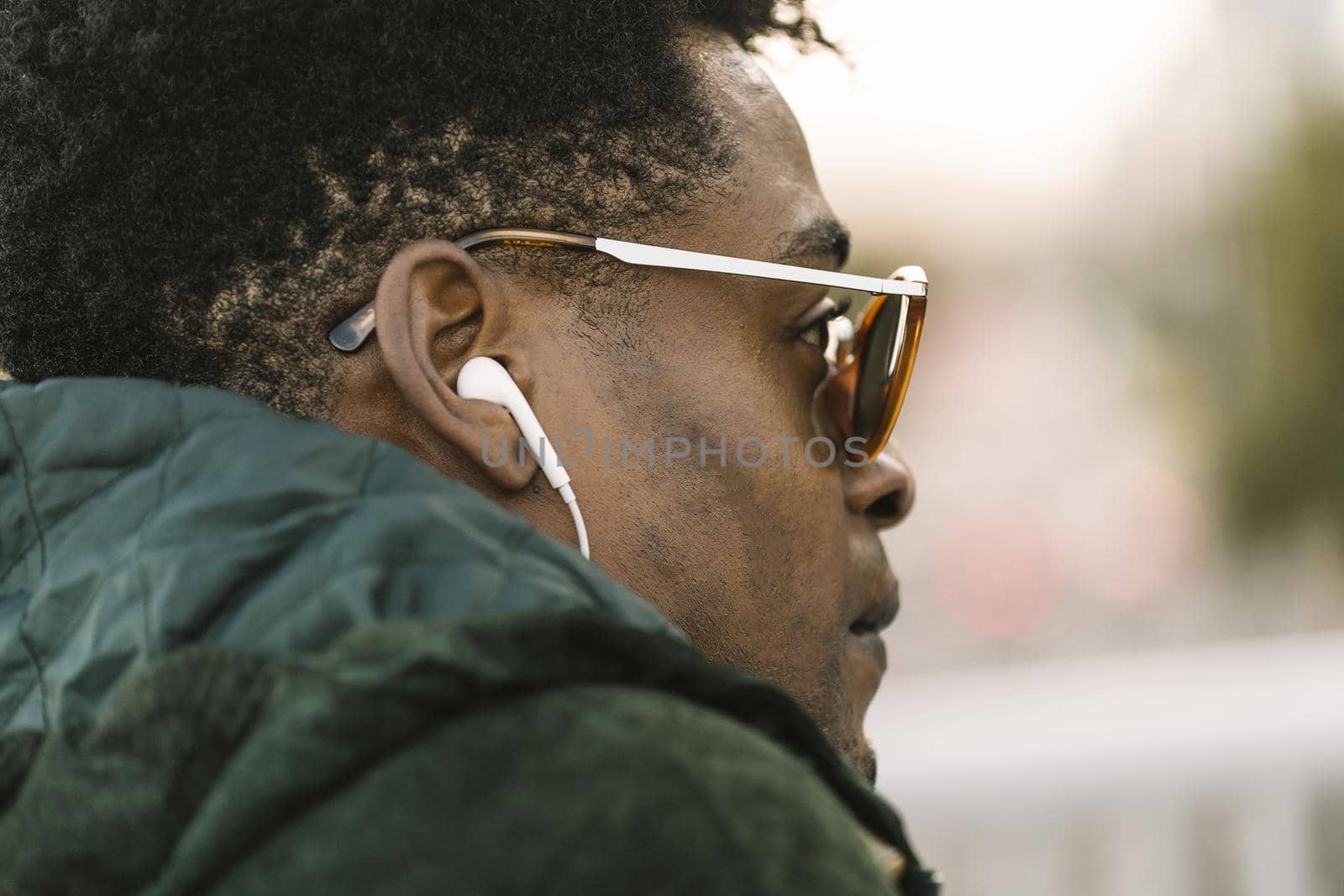 attractive young black african man with green jacket and sunglasses listening music with white earphones, city lifestyle concept