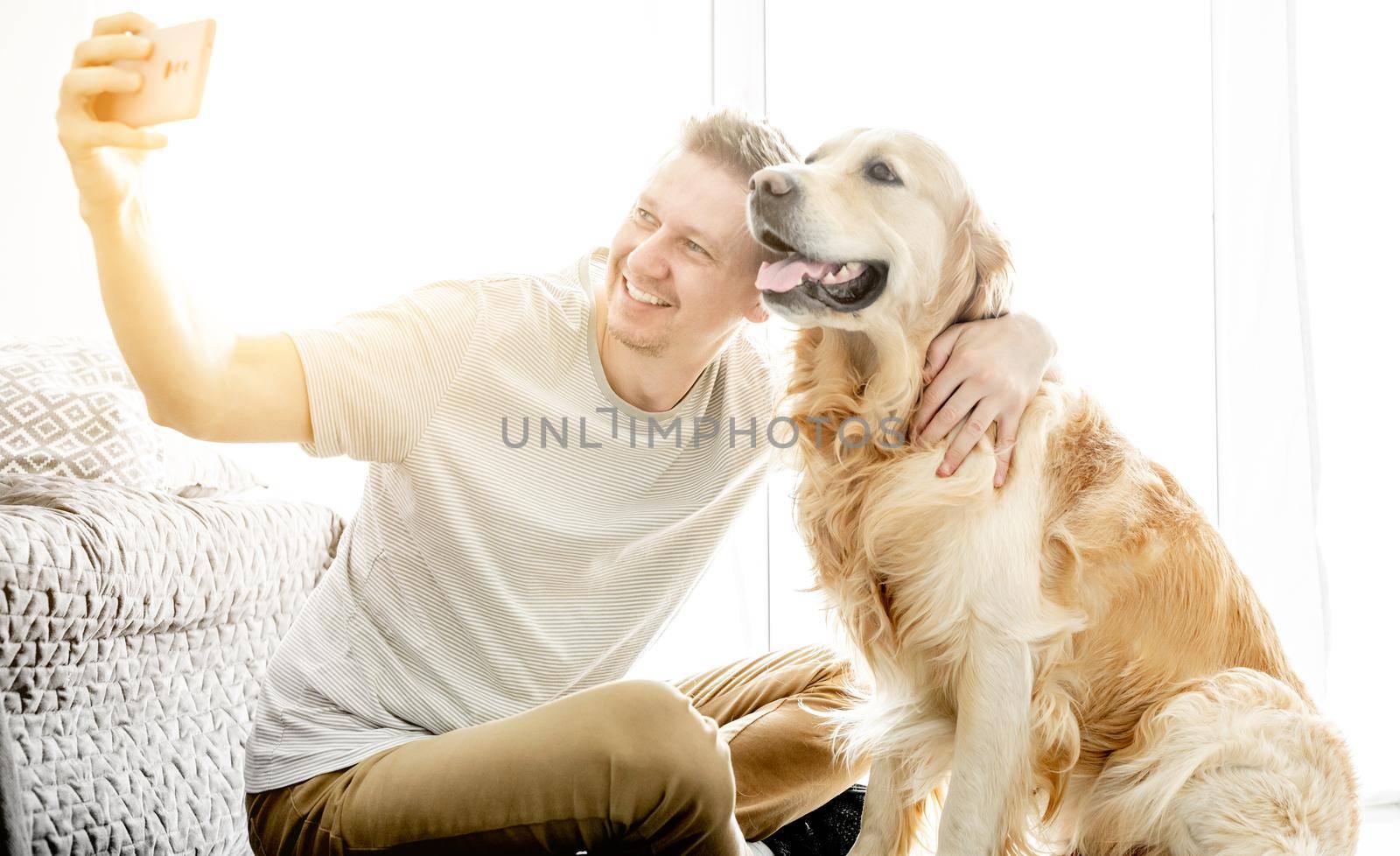 A young handsome man with his golden retriever dog takes a selfie in a bright living room.