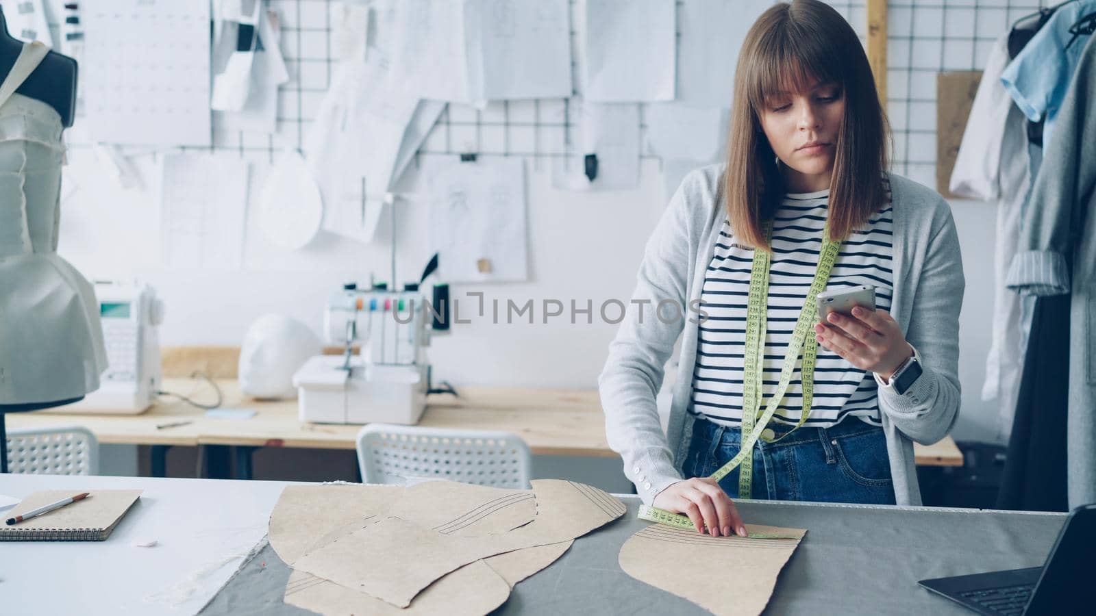 Young seamstress is checking clothing paper patterns and measuring them with tape-measure while looking at smart phone. Nice light studio with garments, textiles and sewing machine in background.