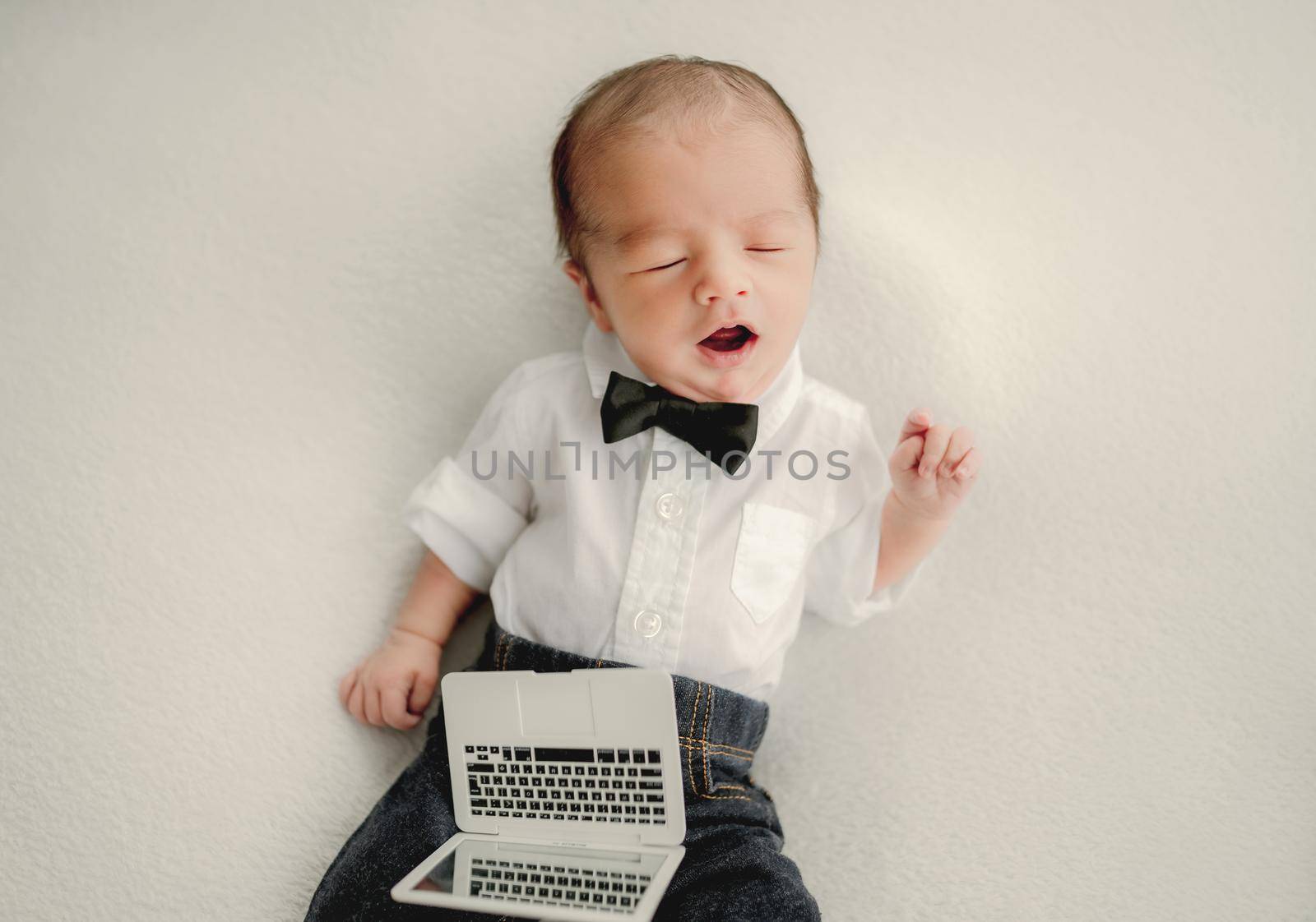 Newborn baby boy wearing festive costume with bow-tie and holding toy laptop falling asleep and yawning. Infant child kid office studio portrait