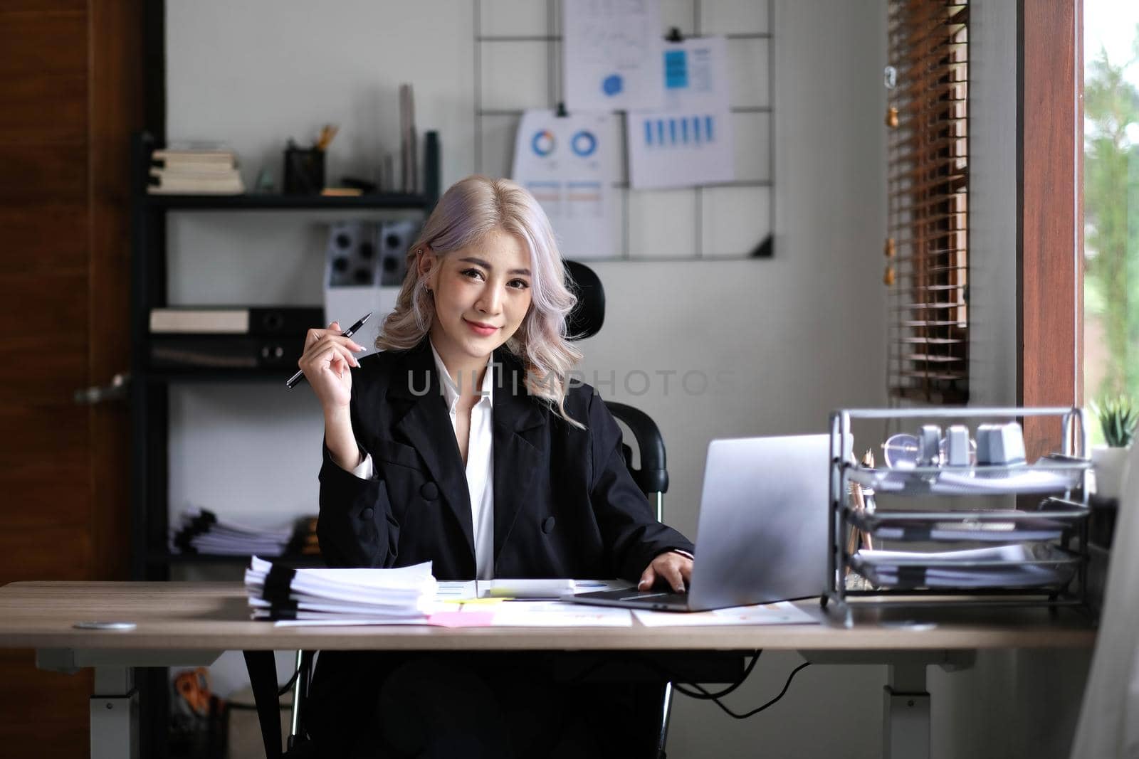 Happy young Asian businesswoman working on laptop keyboard with document at office . Looking at camera..