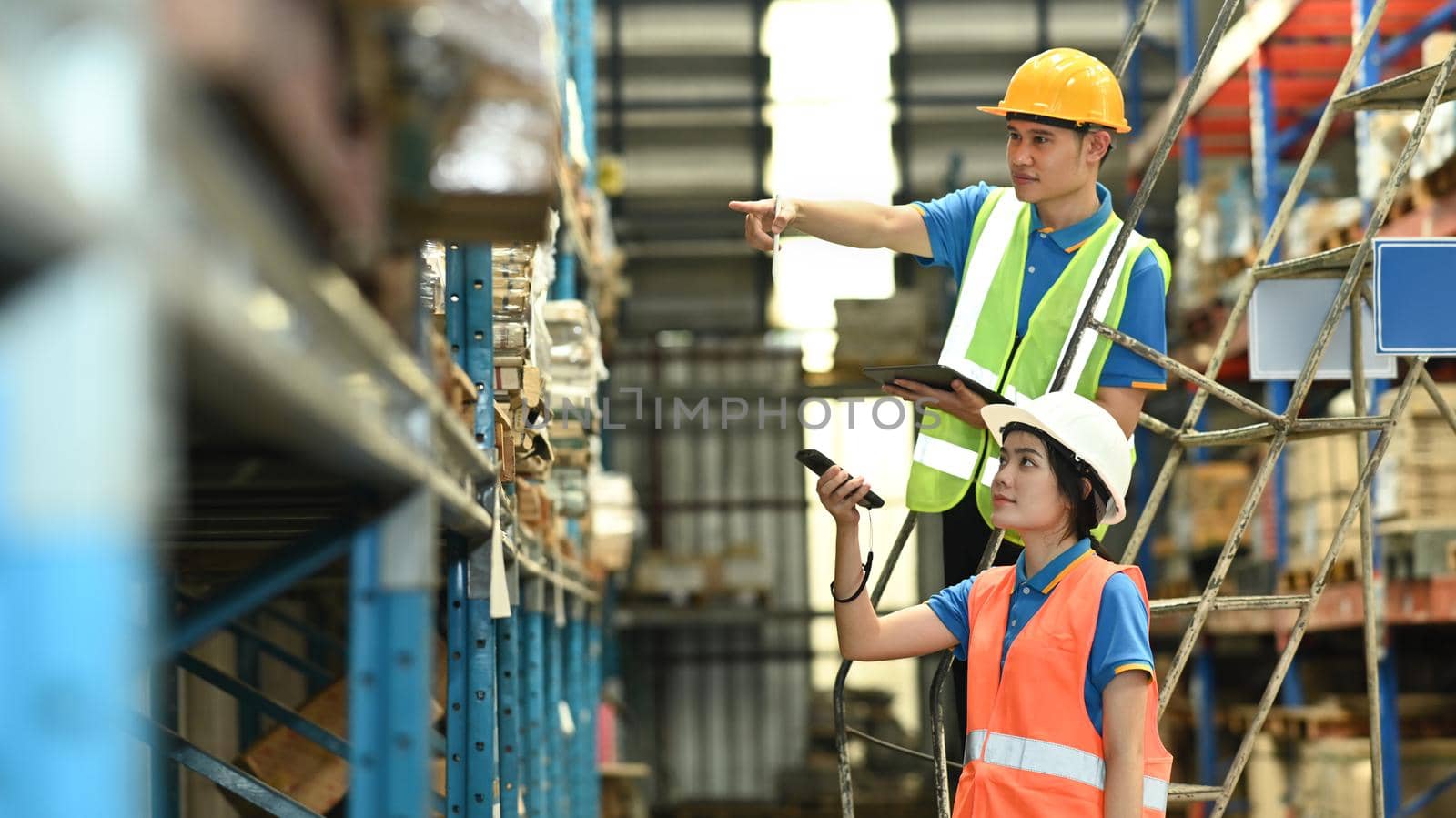 Male and female workers wearing hardhats and reflective jackets checking inventory boxes with barcode scanner in warehouse.