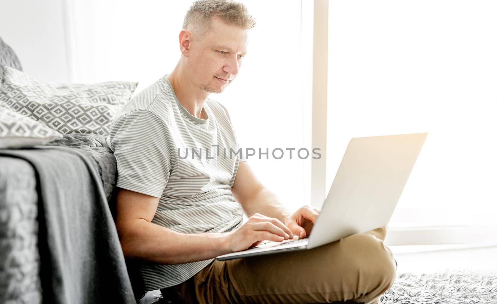 A young handsome man works at a laptop in a bright living room.