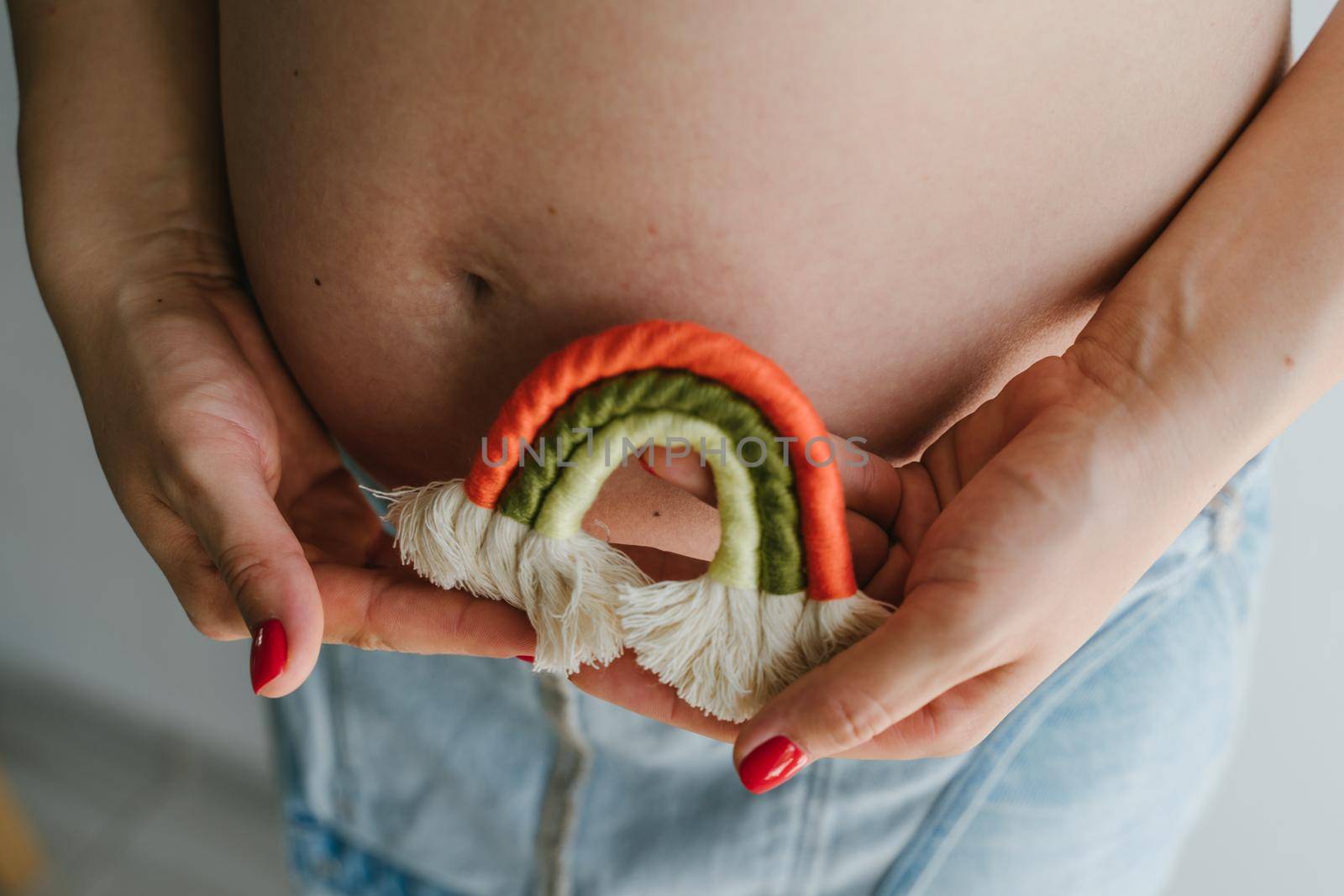 A pregnant girl in a denim jumpsuit holds a rainbow in her hands for the decor of a children's room. Great time to have a baby. Future mom. Rainbow macrame.