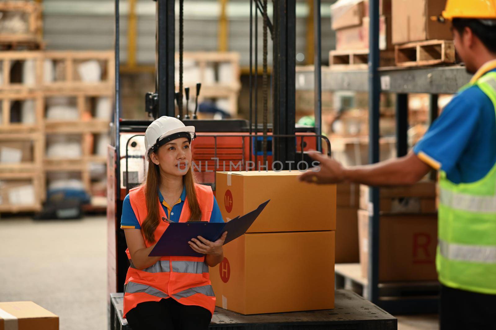 Young female warehouse worker sitting on forklift truck and talking with her colleague.