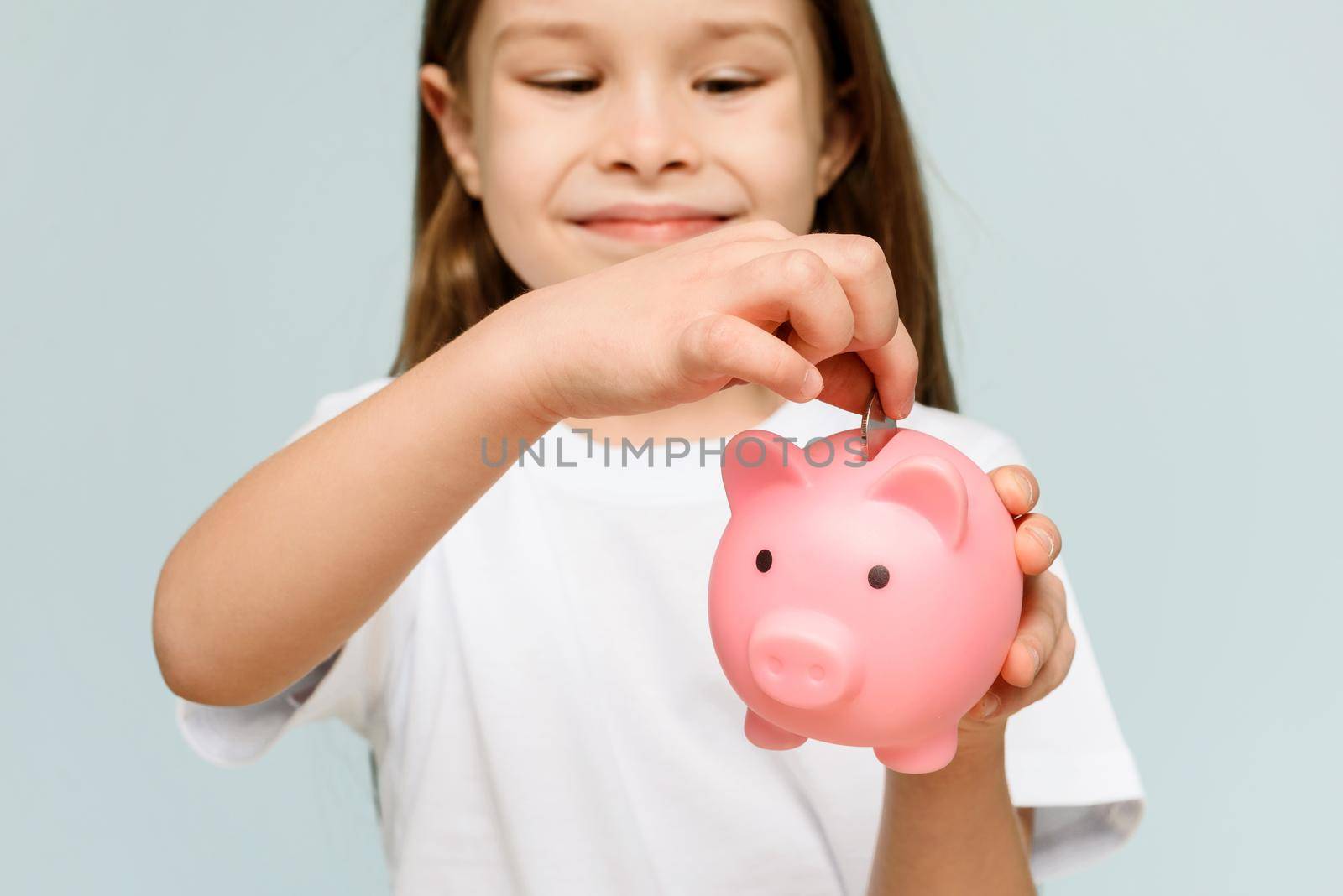 Smiling little girl posing with a piggy bank in her hands, standing against a blue background in a studio with free space. Family savings. by etonastenka