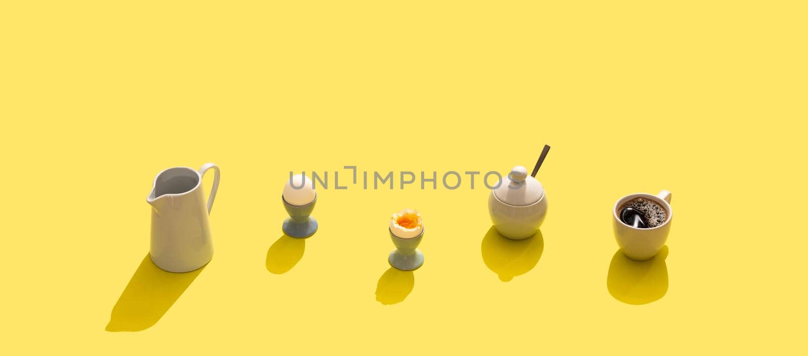 Breakfast set with soft boiled eggs, black coffee in white cup, milk jug, sugar bowl with hard light, yellow bright background. Hard shadows breakfast, simple minimal concept. Copy space, long banner