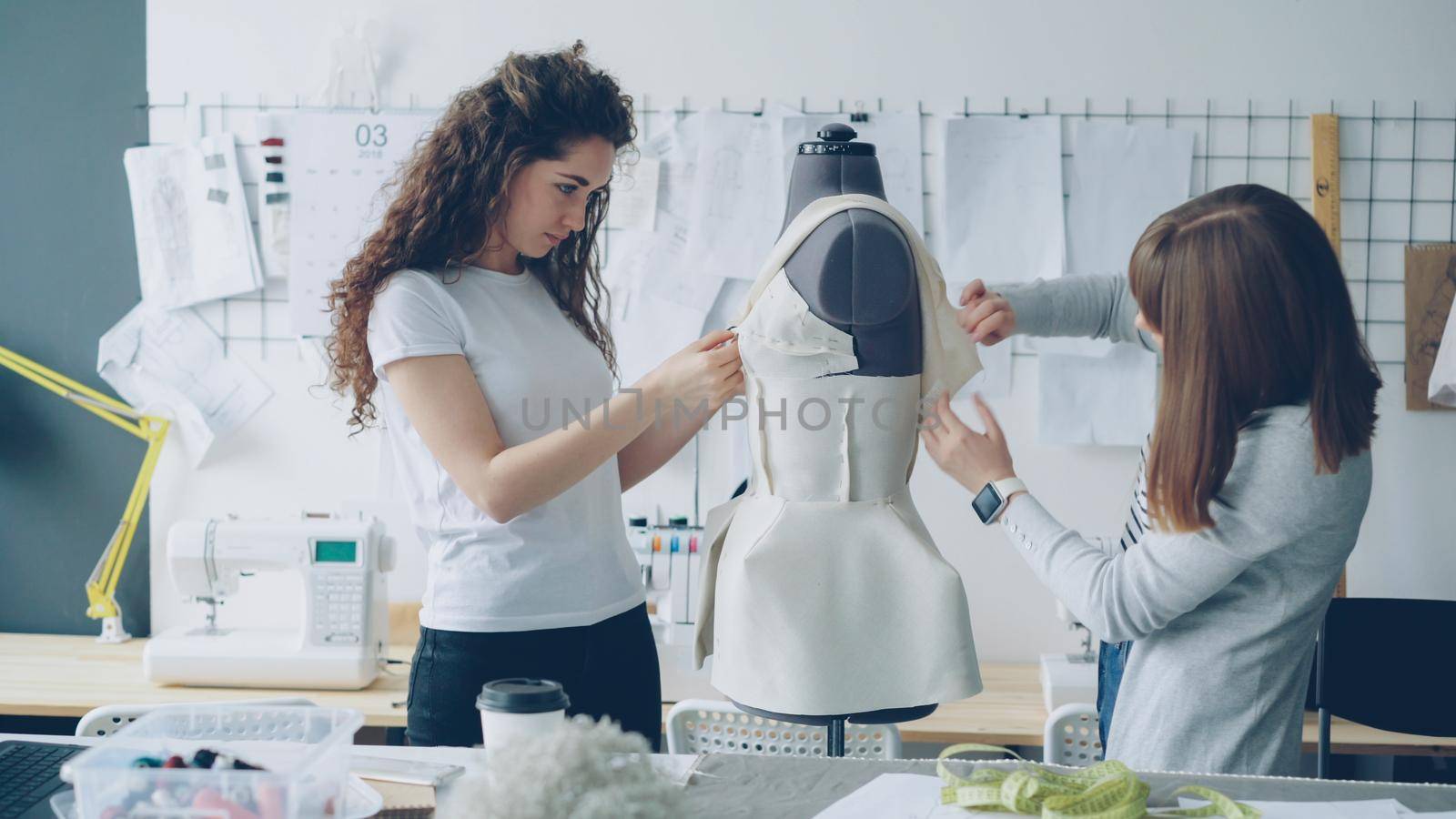 Creative fashion designers are pinning cut out pieces of fabric to mannequin while sewing women's garment in modern studio. Ladies are concentrated on their work. by silverkblack