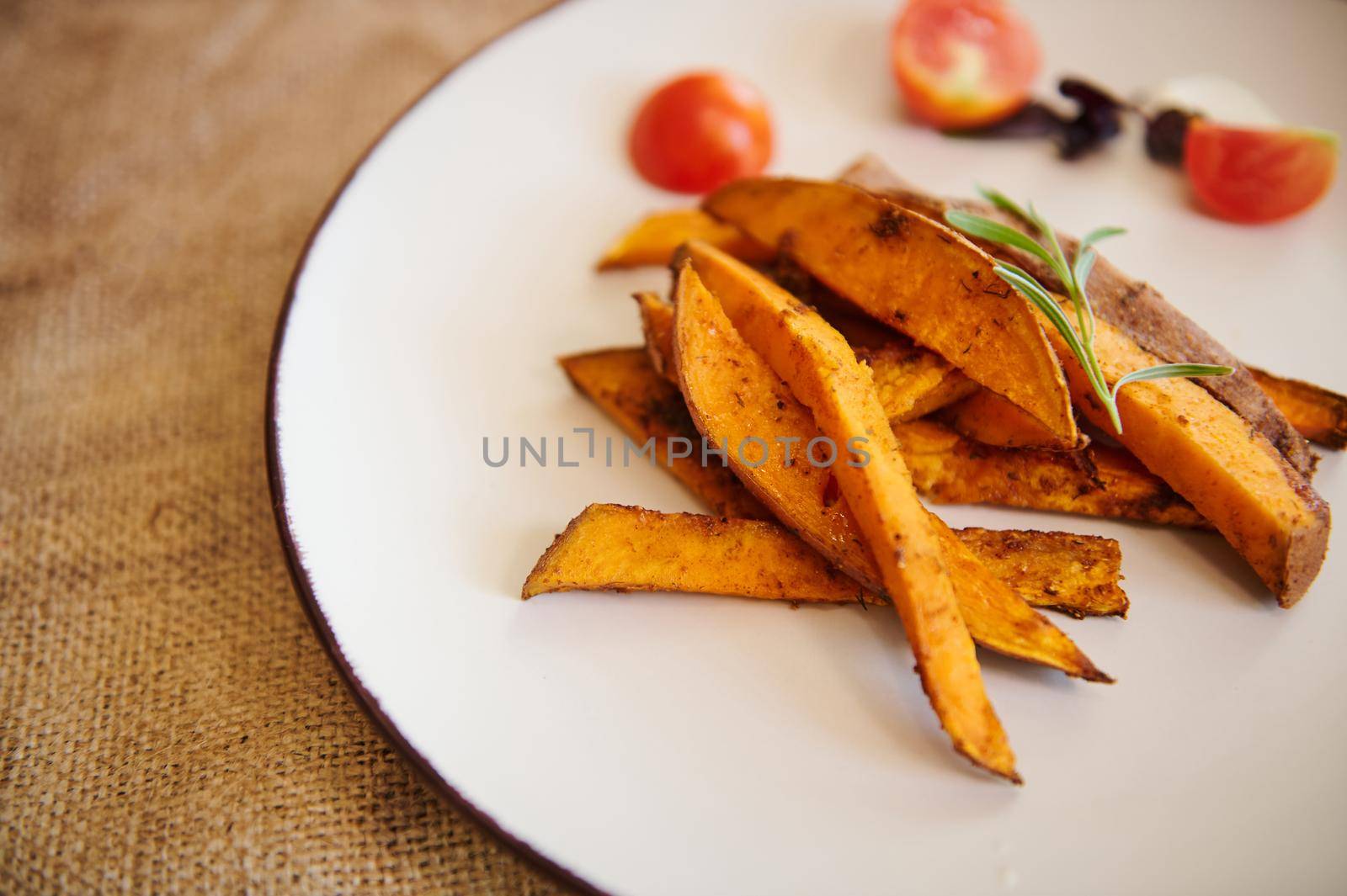 Close-up of a white ceramic plate with wedges of roasted sweet potato with rosemary in a table with linen tablecloth by artgf