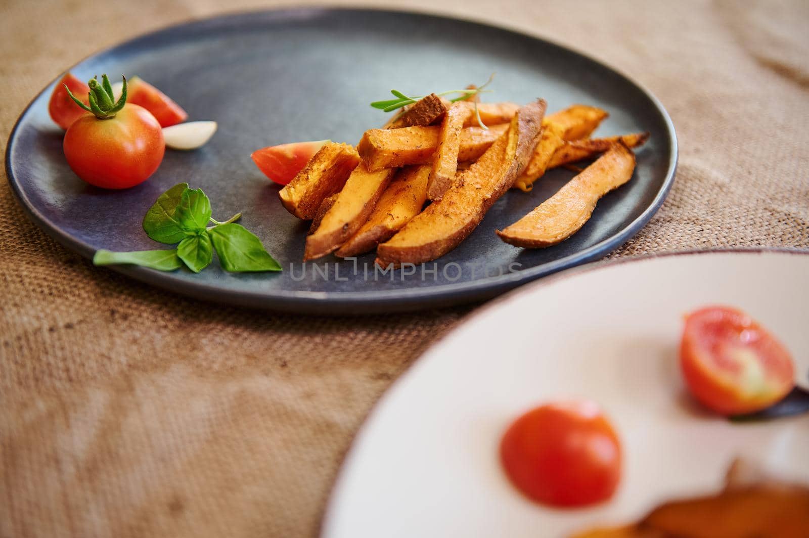 Homemade cooked sweet potatoes with spices and herbs, served with rosemary and cherry tomatoes on blue ceramic plate. Close-up. Roasted batata. Delicious healthy vegan food