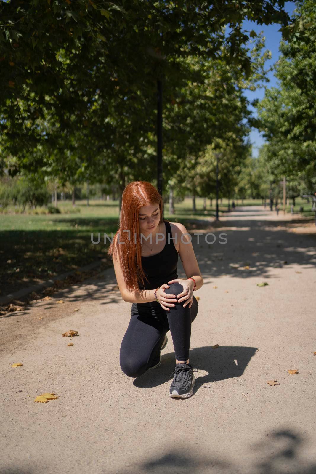 Full body redhead young sportswoman touching dislocated kneecap while kneeling on path during running session on sunny summer day in park