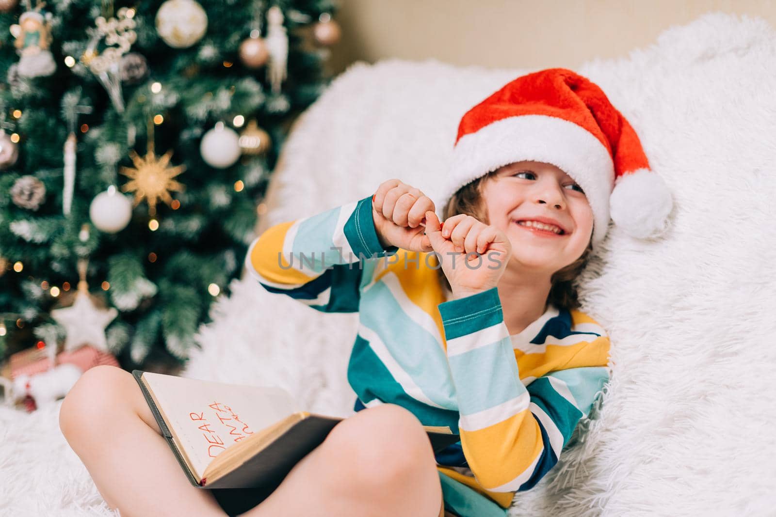 Laughing Kid boy in red Santa hat sit on couch and writing the letter to dear Santa at home. Child wish list. Dreams of a Christmas gifts. Merry Christmas and Happy new year