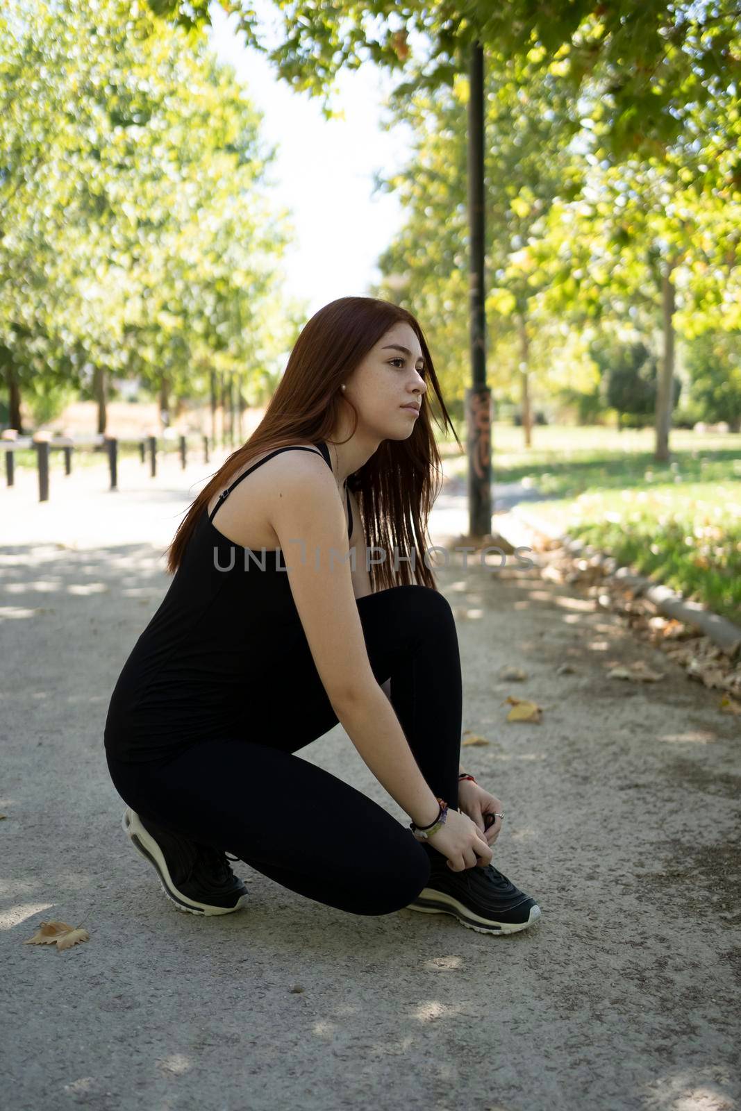 Young modern woman tying running shoes in urban park. by barcielaphoto