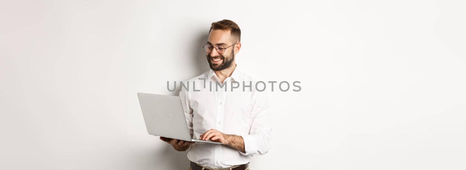 Business. Handsome businessman working on laptop, answering messages and smiling, standing over white background by Benzoix