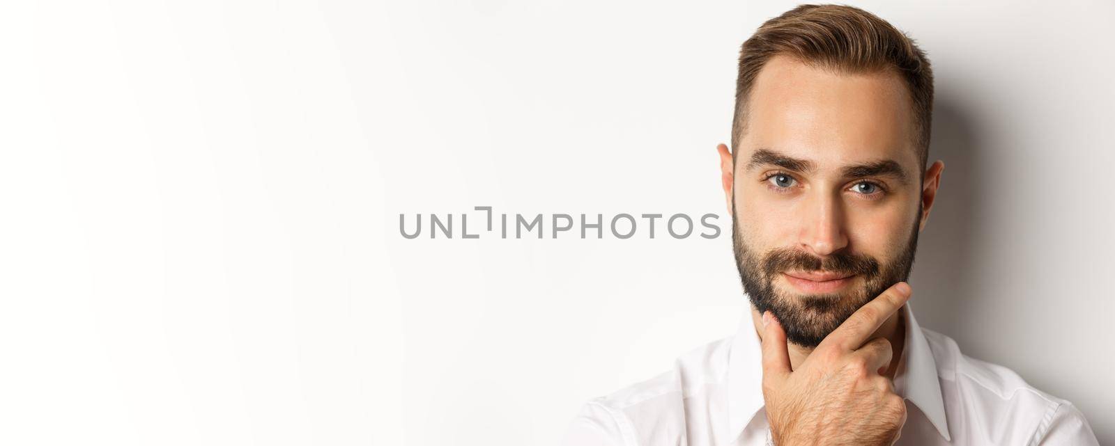 Emotions and people concept. Headshot of handsome thoughtful man smiling satisfied, touching beard and thinking, standing over white background.