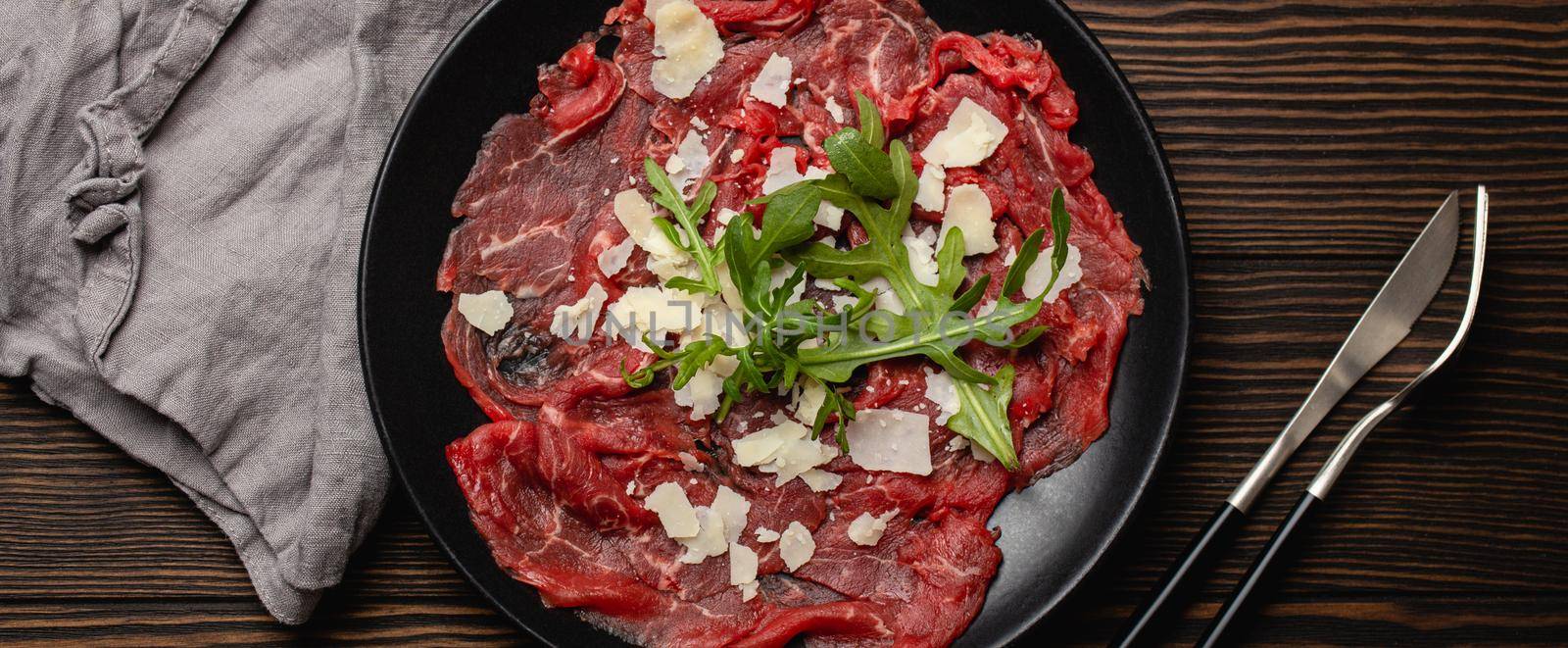 Italian cold meat appetizer Beef carpaccio with parmesan cheese and arugula on black plate with fork and knife on dark brown wooden rustic background flat lay from above .