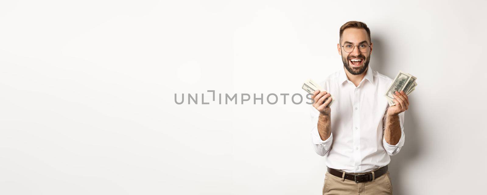 Handsome successful business man counting money, rejoicing and smiling, standing over white background.