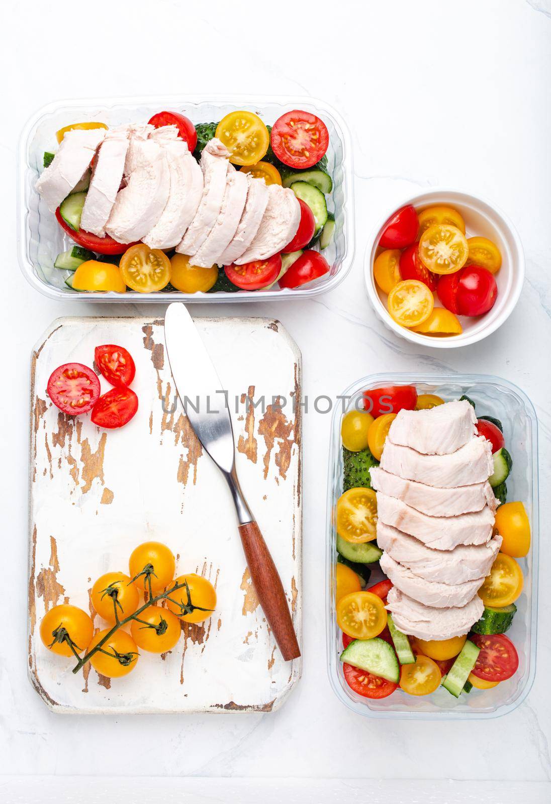 Healthy meal prep for lunch, diet boiled chicken fillet with vegetables salad in lunch boxes containers top view on white marble kitchen table background with knife and cutting board