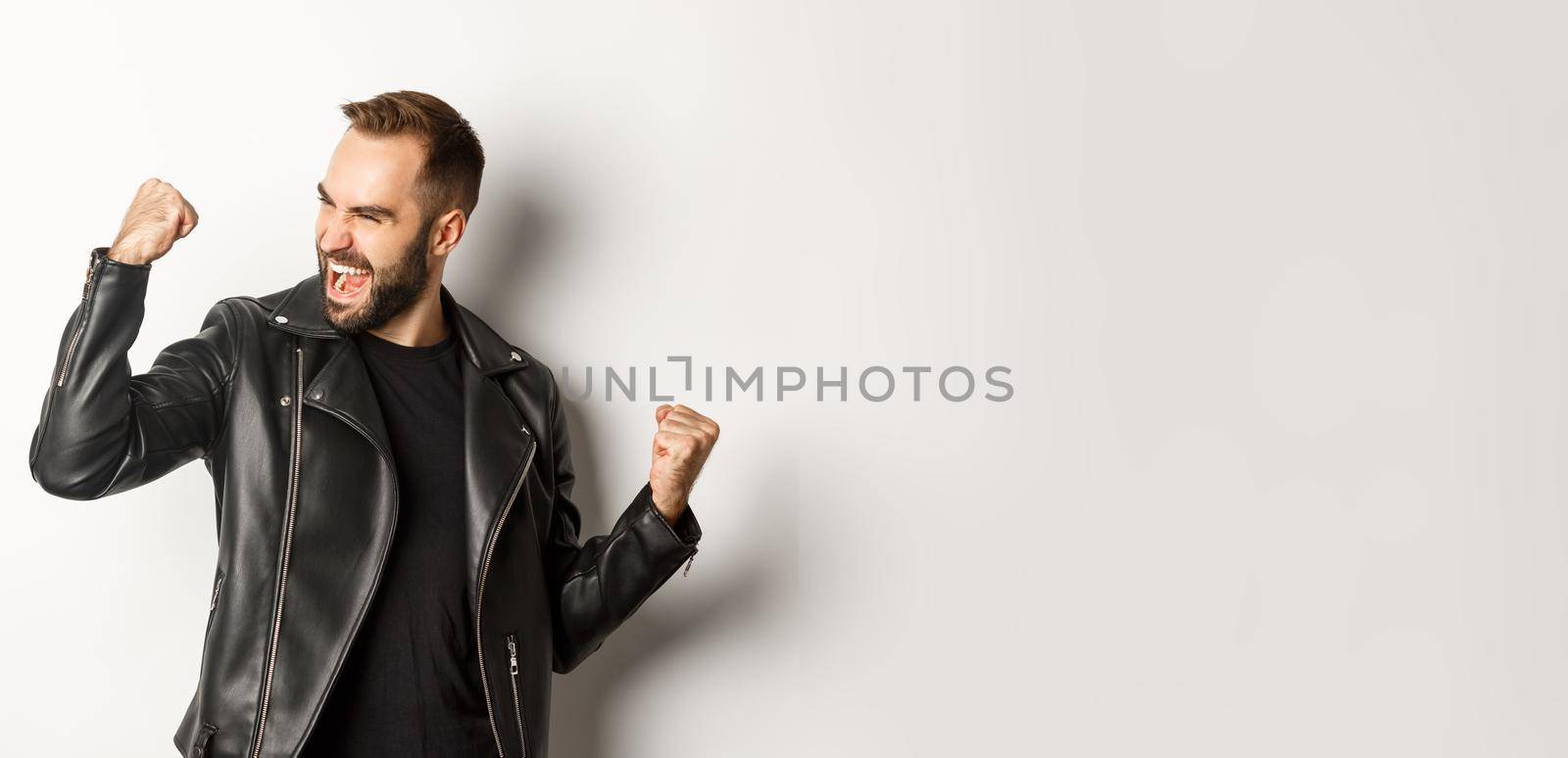 Confident bearded man celebrating victory, winning prize, making fist pump and rejoicing, wearing black leather jacket, white background.