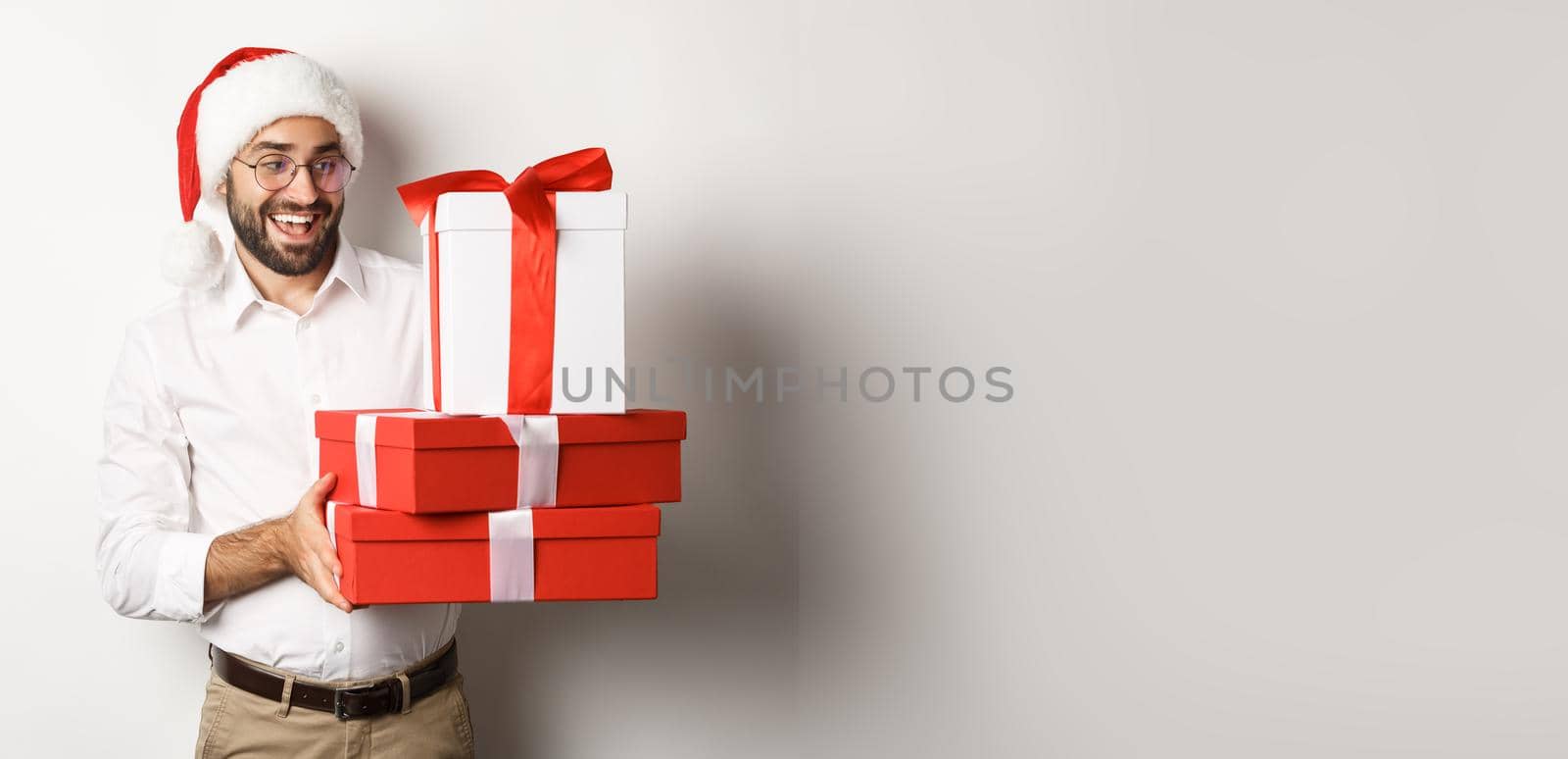 Winter holidays and celebration. Happy guy bring christmas presents, holding gifts and wearing Santa hat, standing over white background by Benzoix